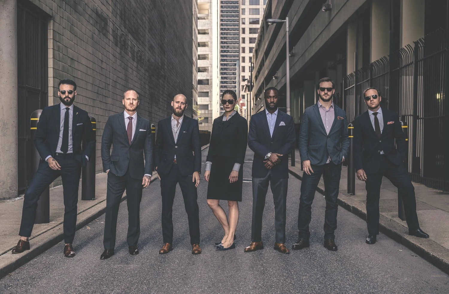 Seven professionally dressed individuals standing in a city alleyway, wearing suits and sunglasses.