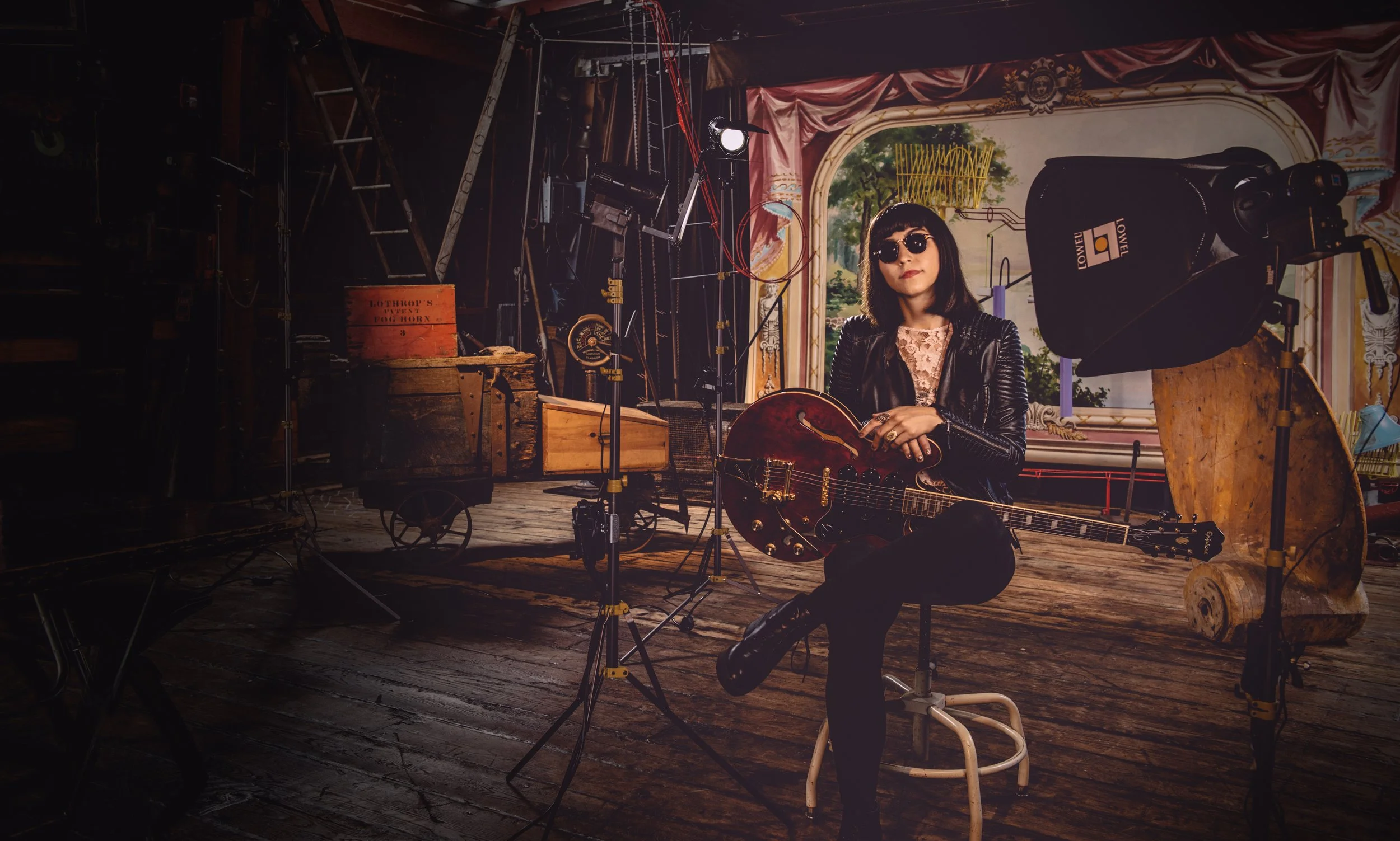 A woman sitting with a guitar in a vintage music studio, surrounded by studio equipment and lighting, with a painted window backdrop of countryside scenery.