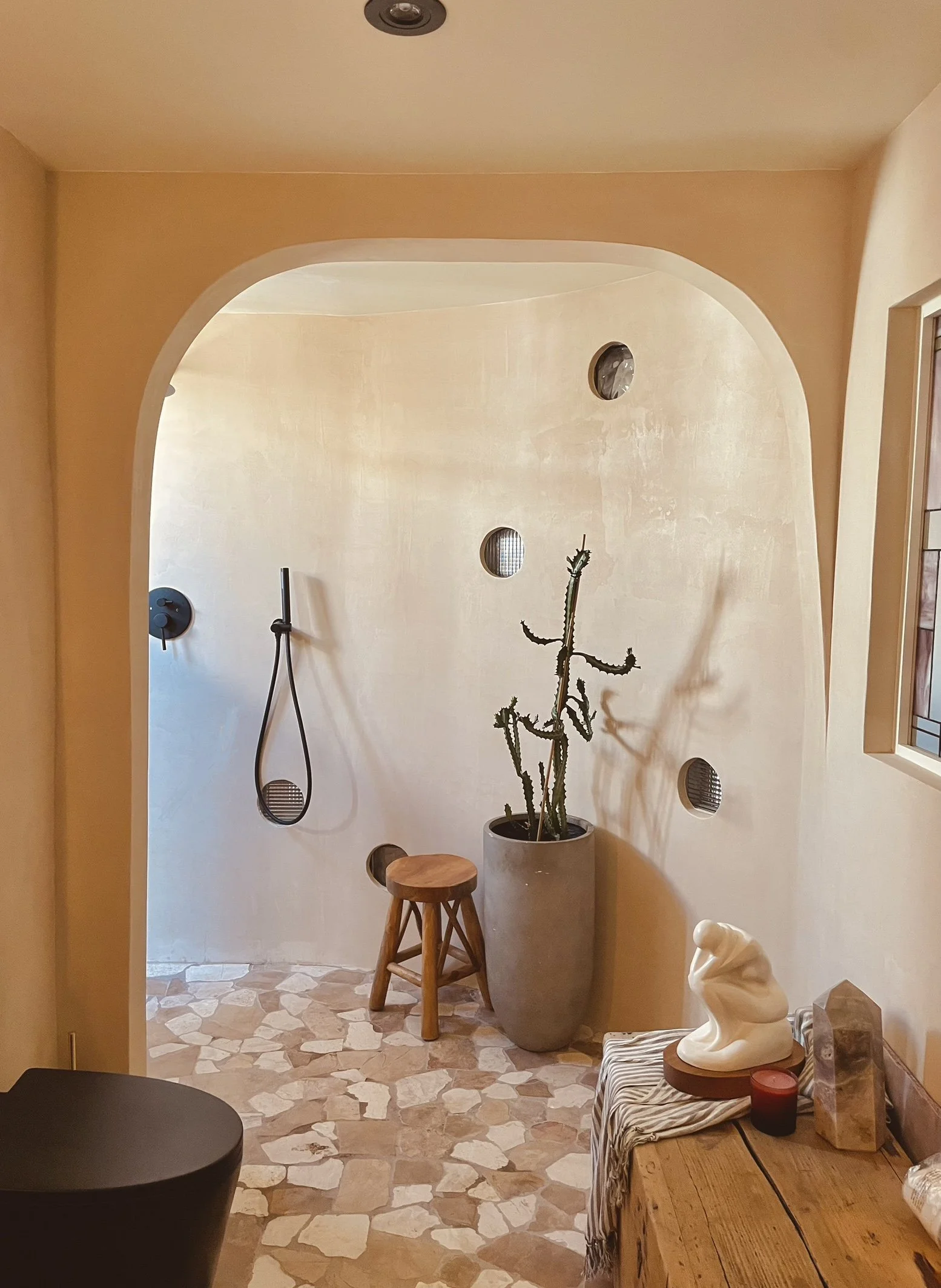 A cozy indoor corner with a tall cactus in a large gray pot, a small wooden stool, decorative wall holes, a sculptural white ceramic piece, a red candle, and a wooden table with a patterned cloth, sunlight streaming through a nearby window.