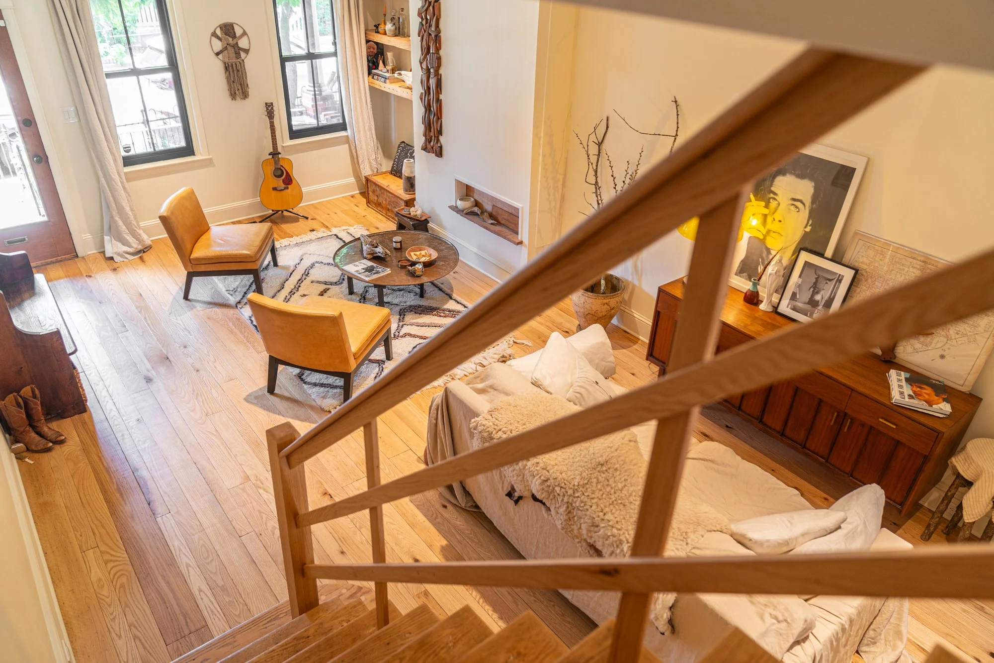 Living room viewed from a staircase, featuring hardwood floors, a tan sofa with throw blankets, a wooden coffee table, two tan chairs, a guitar near windows, decorative artwork, and a wooden sideboard with framed pictures and a lamp.
