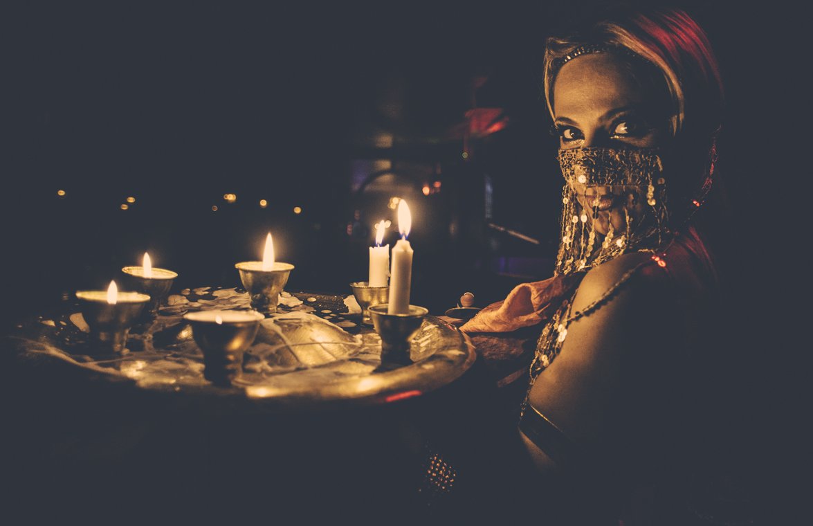A woman with red hair wearing a beaded face covering and a sleeveless top, sitting at a table with lit candles in an otherwise dark environment.