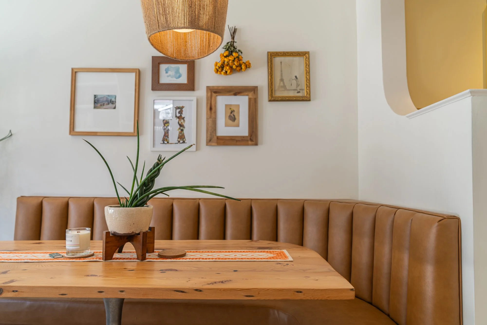 Interior of a cozy cafe or dining area featuring a wooden table with a potted plant, a candle, and a remote control. The background includes a white wall with a gallery of framed artwork and a hanging light fixture.