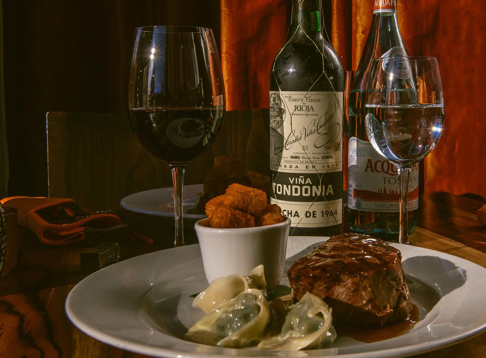 A dinner scene with a glass of red wine, a glass of water, a plate of meat with sauce, a small bowl of fried foods, and a small bowl of cheese with dough. There are two bottles of wine in the background.