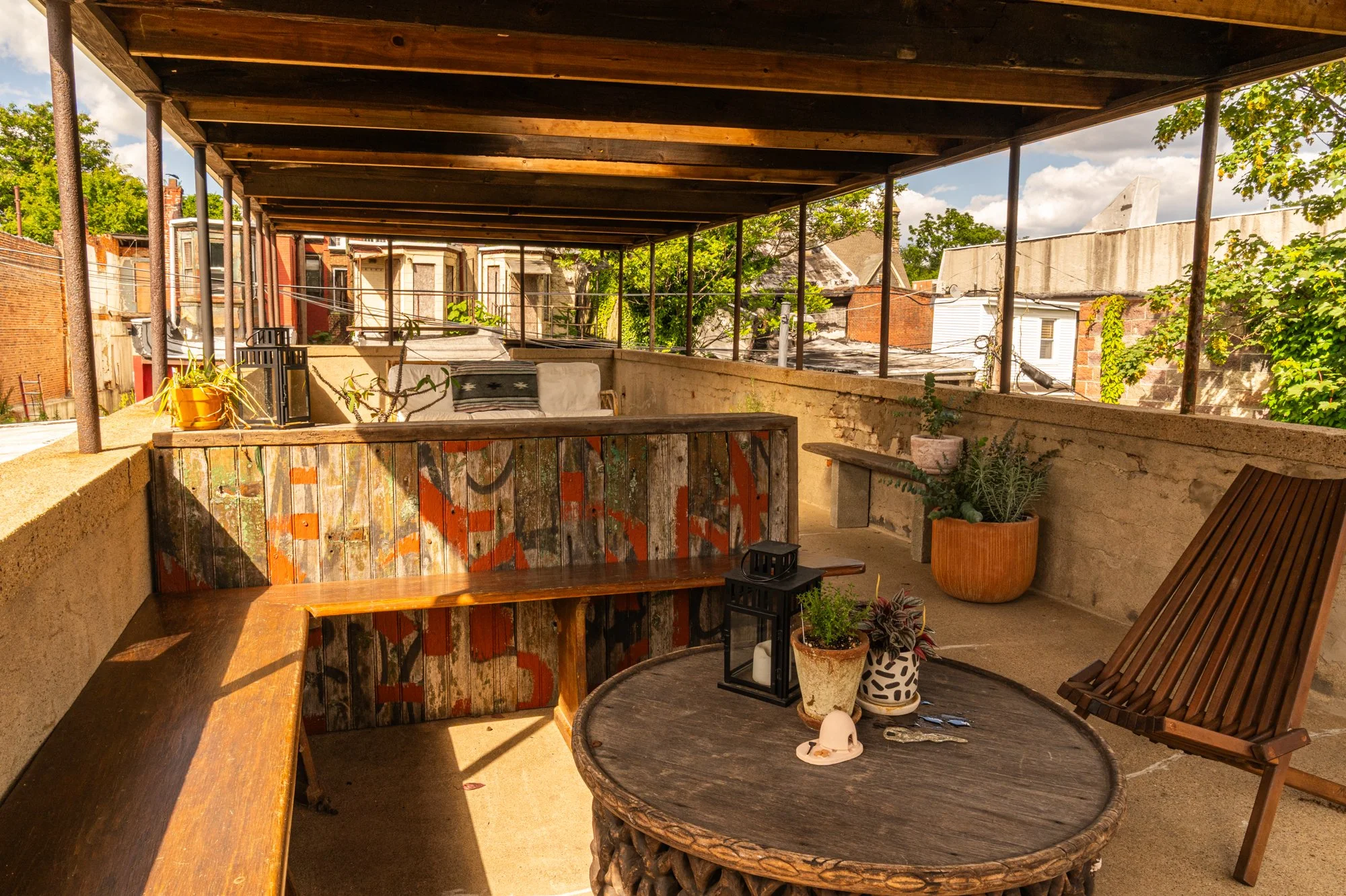 A cozy rooftop patio with wooden furniture, potted plants, and a mix of rustic and modern decor, overlooking neighboring buildings and partly cloudy sky.