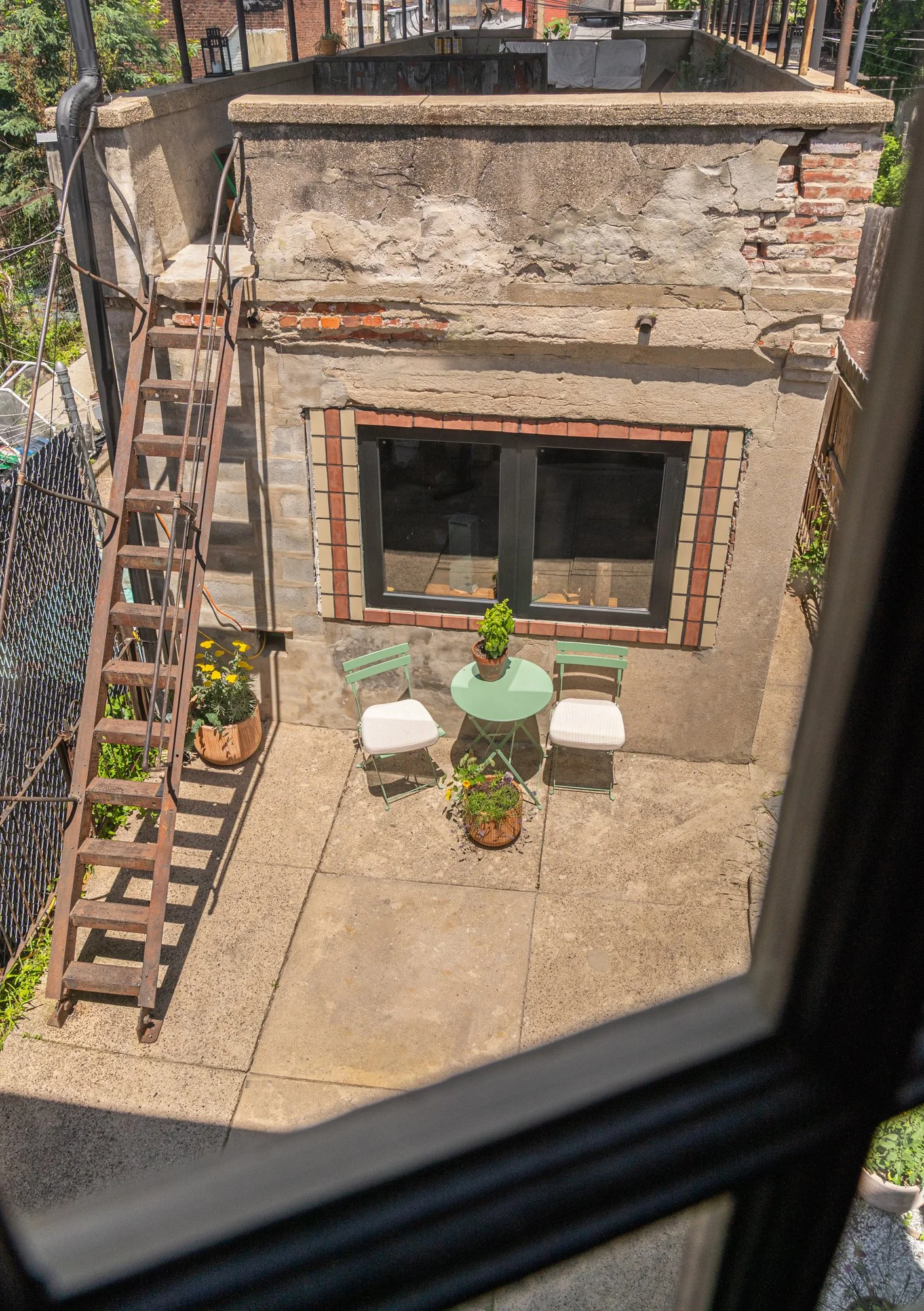 View of a small outdoor patio or backyard seen from a second-story window. The patio has a concrete floor, a green round table with a potted plant on top, two white chairs with green frames, and a small potted plant on the ground. A brick building wa