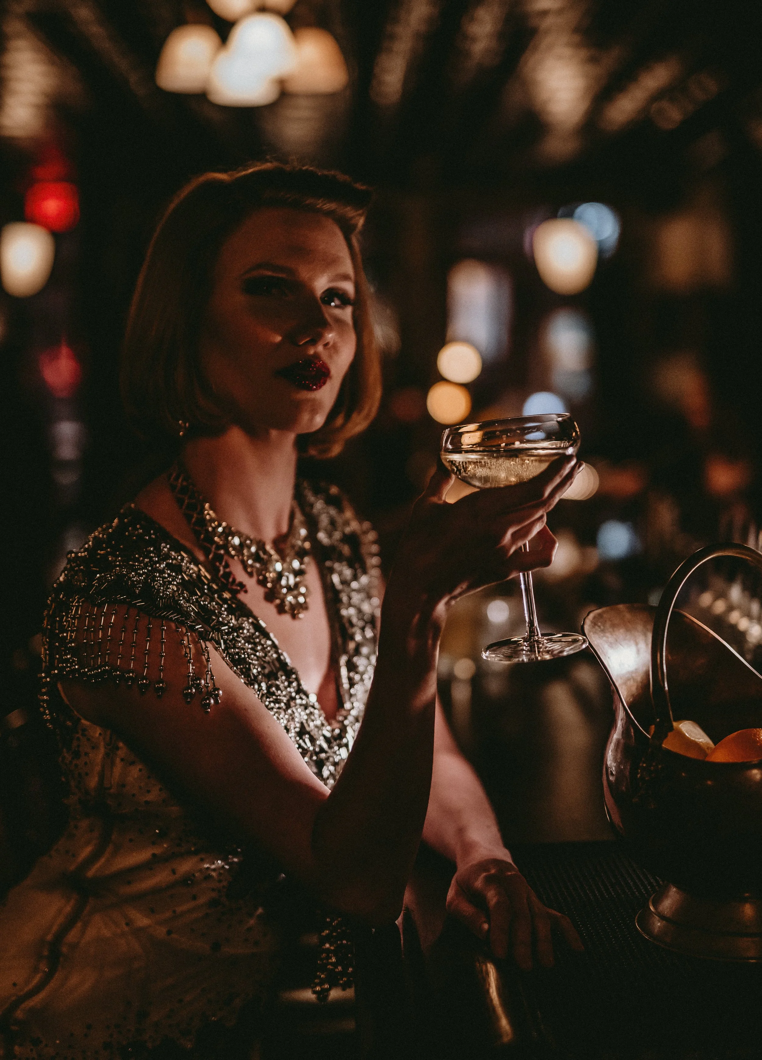 A woman in an embellished dress holding a cocktail glass at a dimly lit bar or club, with blurred background lights.
