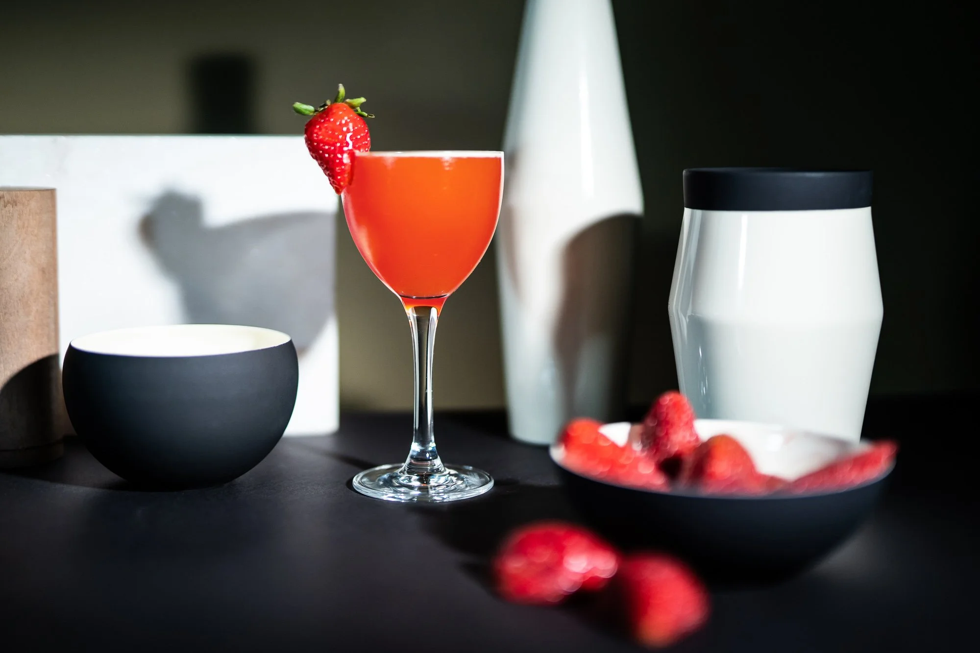 A glass of pink cocktail with a strawberry garnish, black and white bowls and containers, and fresh strawberries on a black surface.