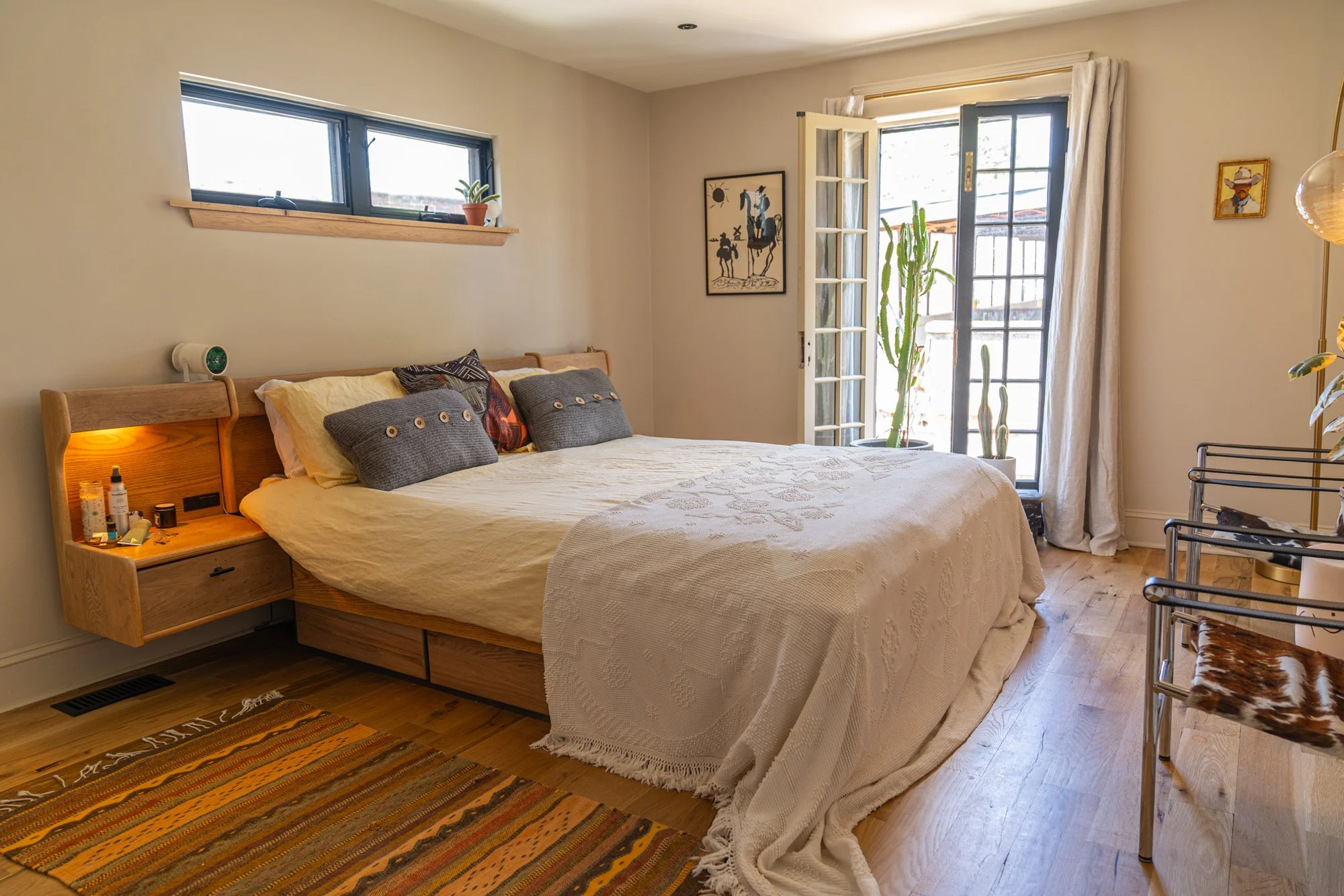 A cozy bedroom with a wooden bed frame, white bedding, and throw pillows, next to a windows and a glass door with white curtains, featuring potted cacti and wall art.