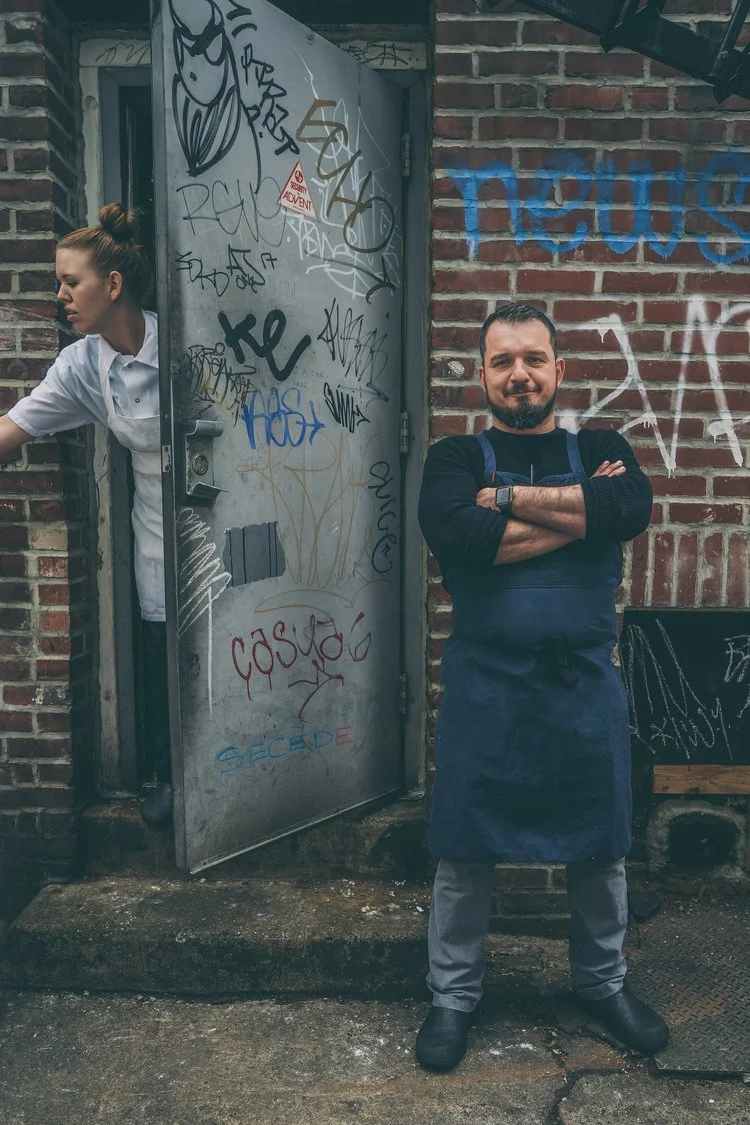 Man standing with arms crossed in front of a graffiti-covered brick wall and door, woman in white shirt behind door, graffiti including tags and drawings.