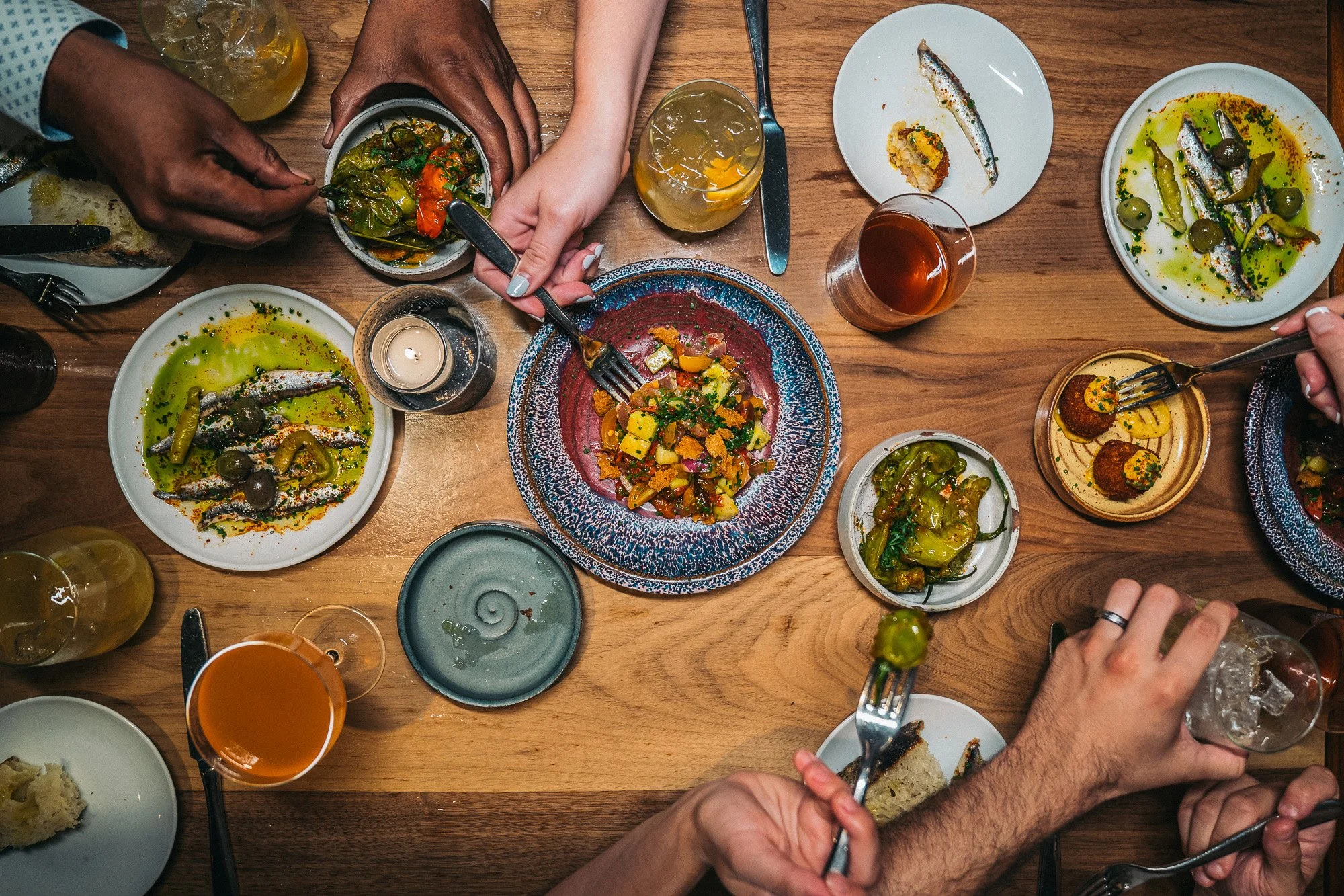 People sharing a meal at a wooden dining table with various plates of food, drinks, and utensils, viewed from above.
