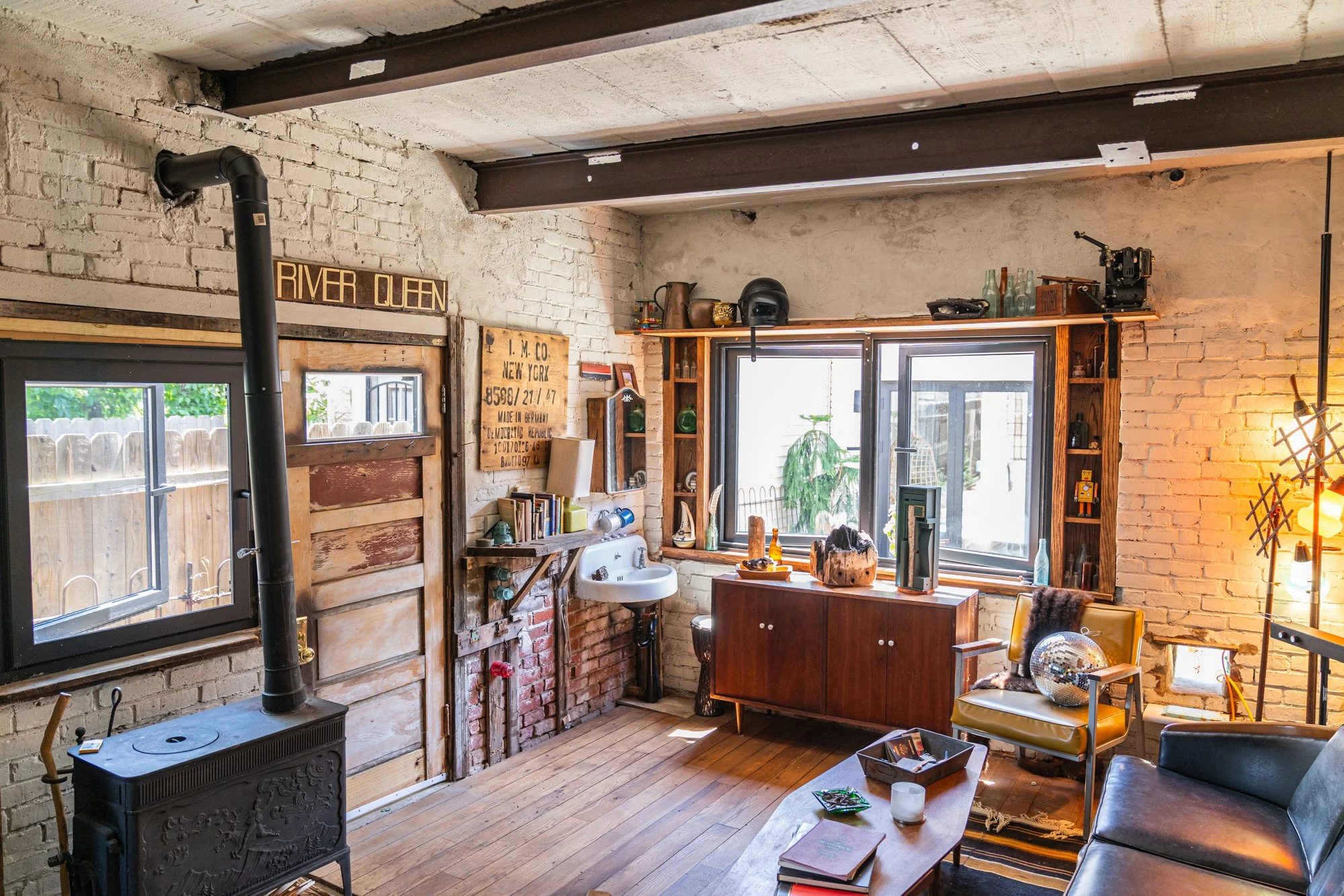 Cozy living room with brick walls, wooden floors, and large windows. Contains a vintage wood stove, a small sink, a yellow armchair with a disco ball, a black sofa, a wooden coffee table with books and a candle, a sideboard with decorative items, and