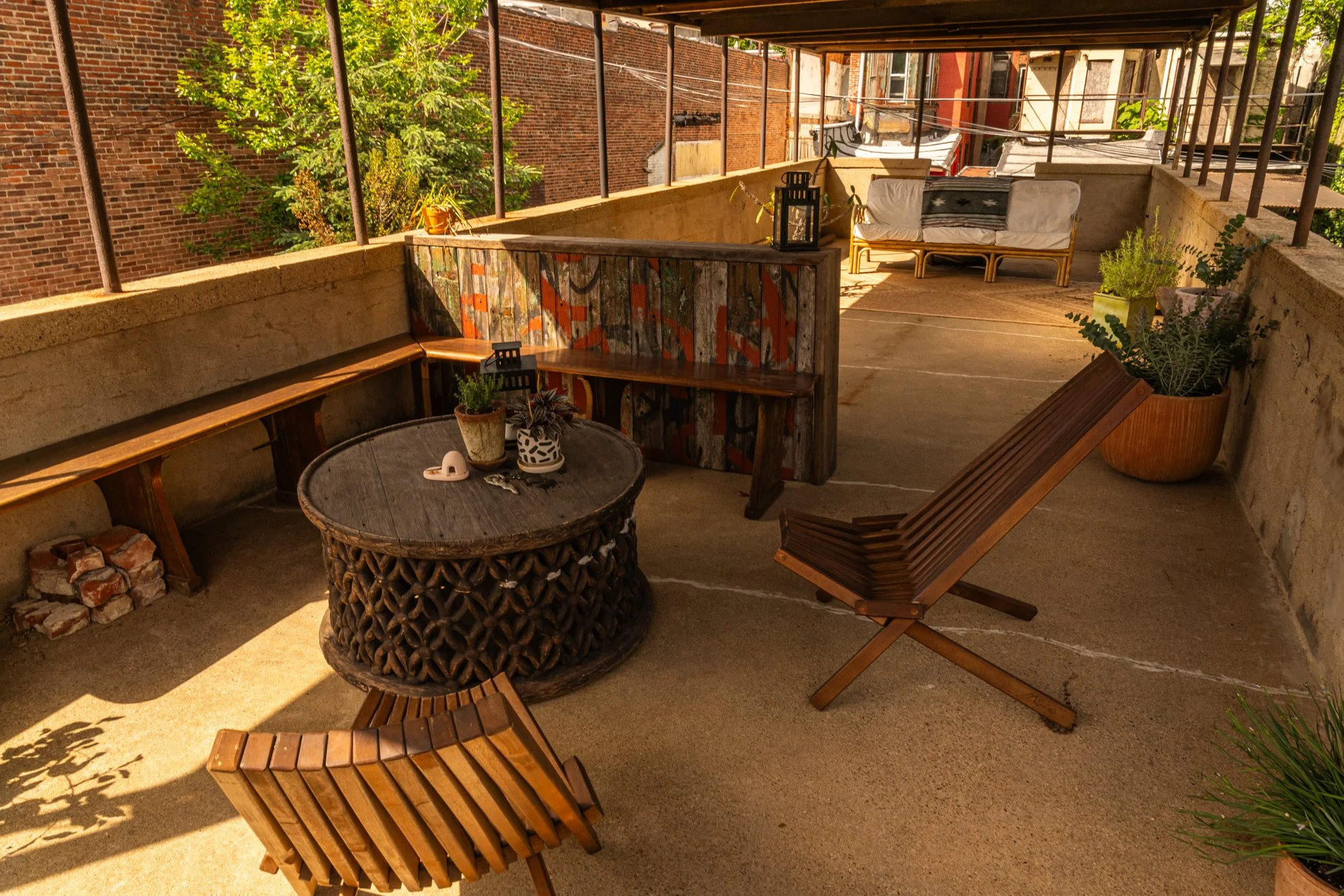 An outdoor balcony with wooden furniture, potted plants, a colorful painted wooden partition, and a stone table with decorative items on it, surrounded by brick buildings and trees.