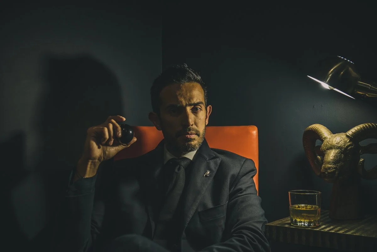 Man in a dark suit sitting in a dimly lit room, holding a cigar, with a glass of whiskey and a decorative ram's head sculpture on a side table.