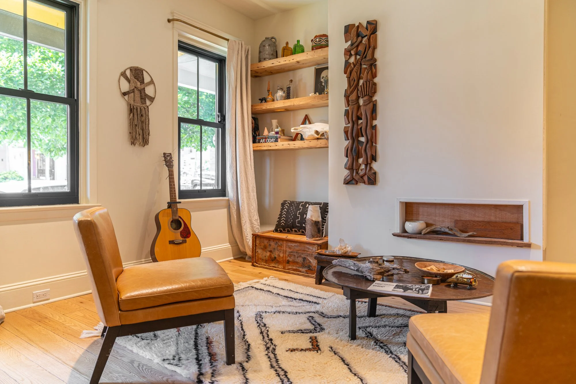 Cozy living room with two tan chairs, a guitar near the window, a round coffee table with decorative items, and a white rug with black and orange accents. Two large windows with black frames and white curtains let in natural light. Wooden shelves hol