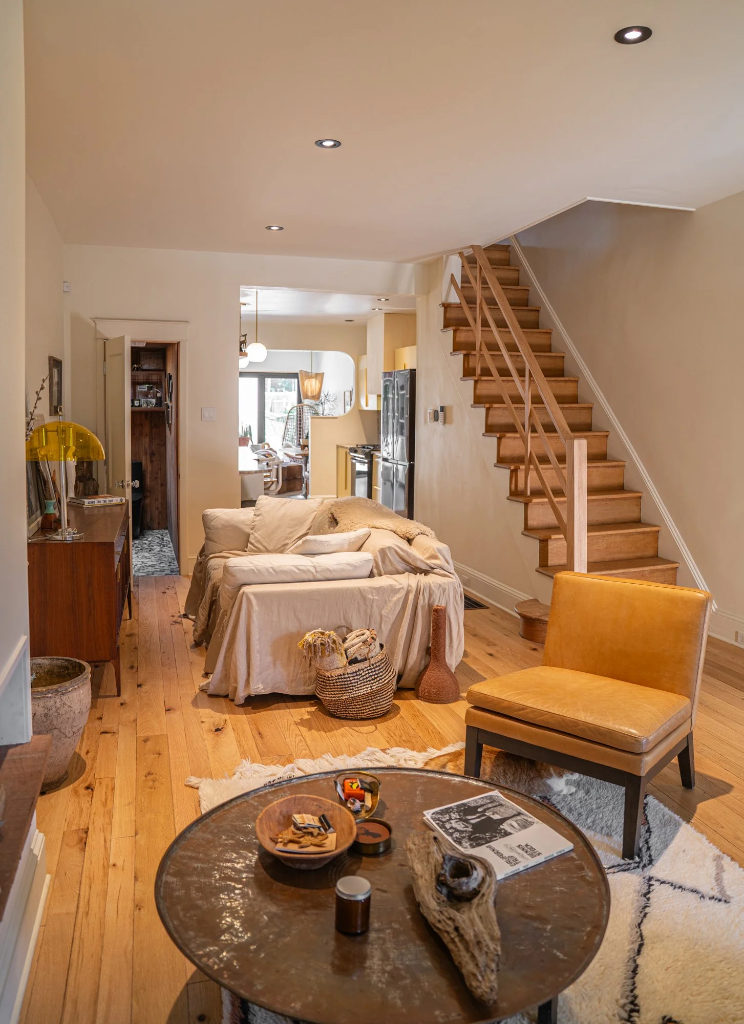 Living room with wooden floors, beige walls, and a sofa covered with a beige cloth. There is a small coffee table with various items including a magazine, a bowl, and a candle. A mustard yellow chair is nearby. A staircase with wooden steps and a min