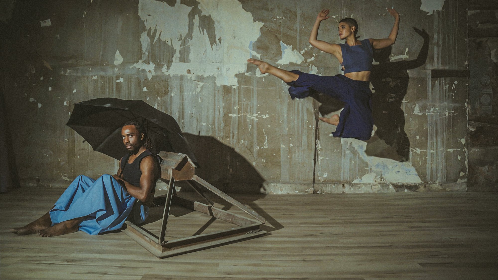 A woman in a blue crop top and skirt is airborne, performing a kick in a dance studio with a distressed wall. A man with dreadlocks sits on the floor, holding a black umbrella and watching her.
