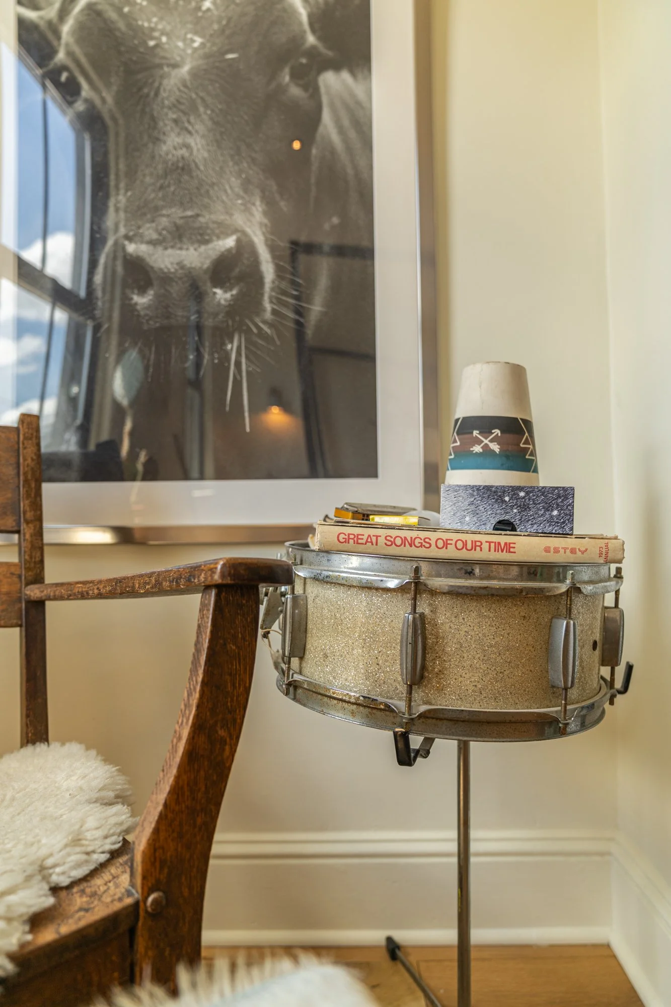 Close-up of a stereo drum with magazines, a cup, and small objects on top, next to a wooden chair with sheepskin, and a large black-and-white photo of a dog's face on the wall.