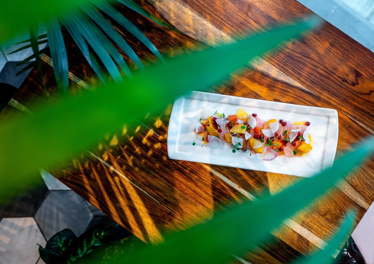A rectangular white plate with a colorful salad on a polished wooden table seen through green leaves in a bright setting.