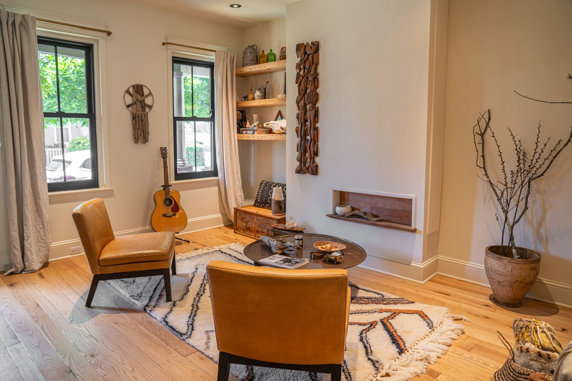 Cozy living room with two beige chairs around a wooden coffee table on a black and white patterned rug, guitar by window, wooden shelves with decor items, large potted plant, and natural light coming through two windows.