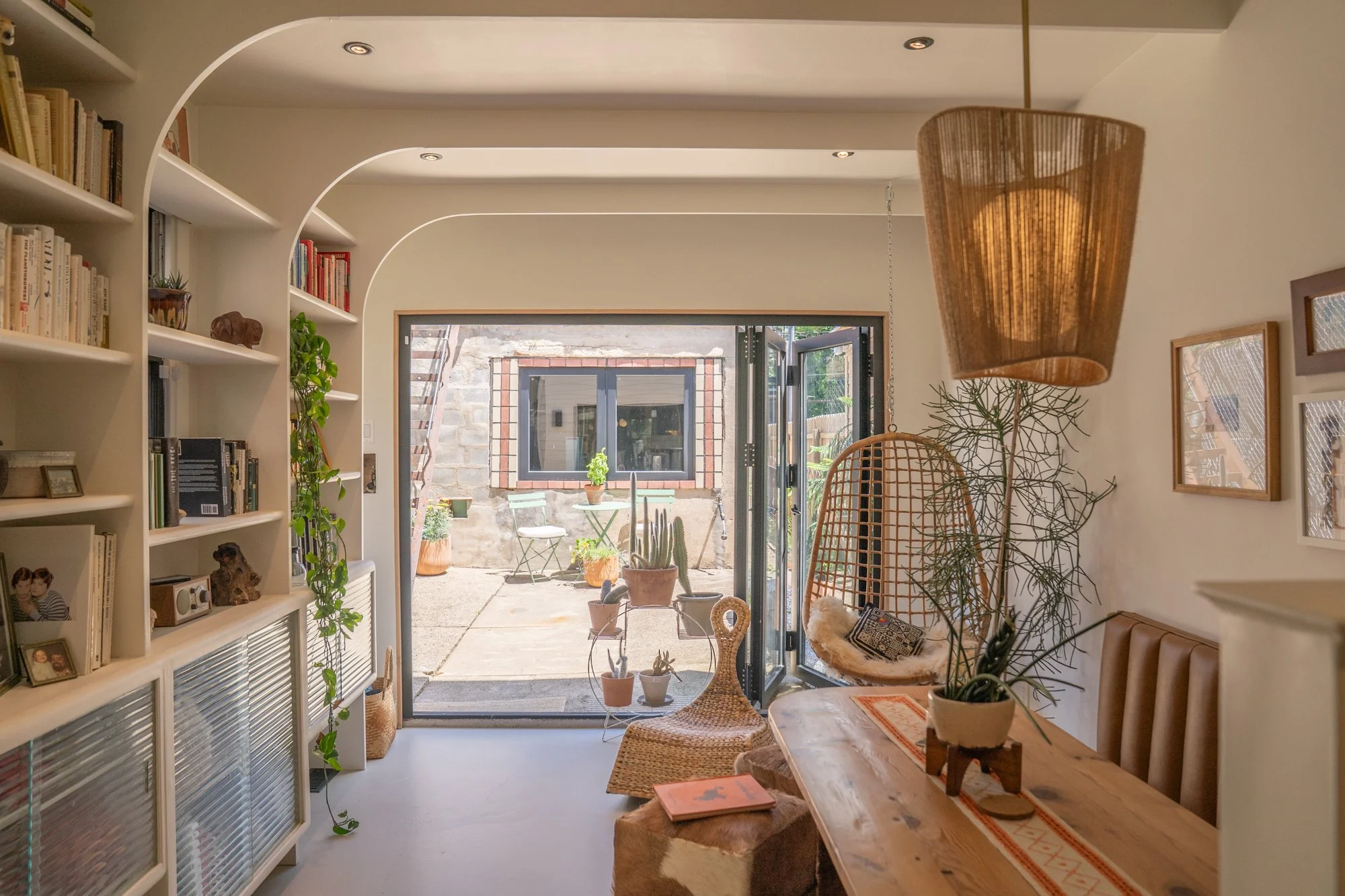 Interior of a cozy room with a wooden dining table, surrounded by wicker chairs and a hanging lamp. The room opens to a patio with potted plants and a small table with chairs outside.