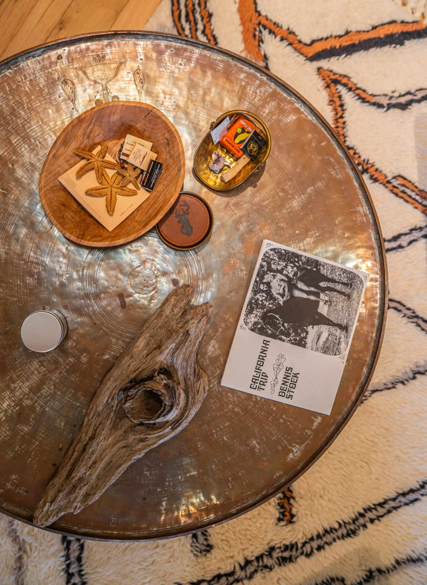 Top-down view of a round metal table with various decorative items including a wooden driftwood piece, a paper pamphlet with a black and white photo, starfish, a small round container, a leather patch, and a small bowl with matchboxes and a lighter. 