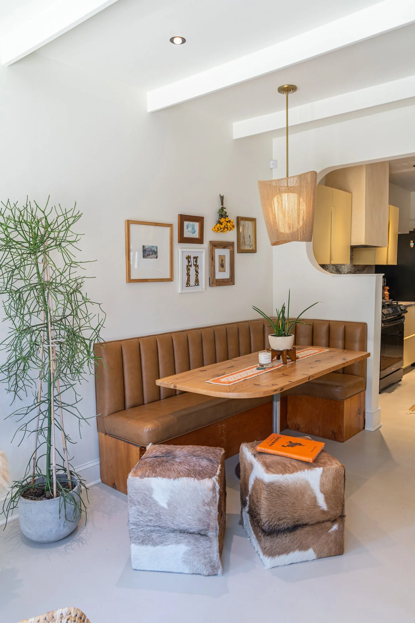 Cozy dining nook with a tan leather bench, a wooden table, two cowhide ottomans, and a small potted plant. Wall decor includes framed pictures and hanging flowers, with a woven pendant light overhead.