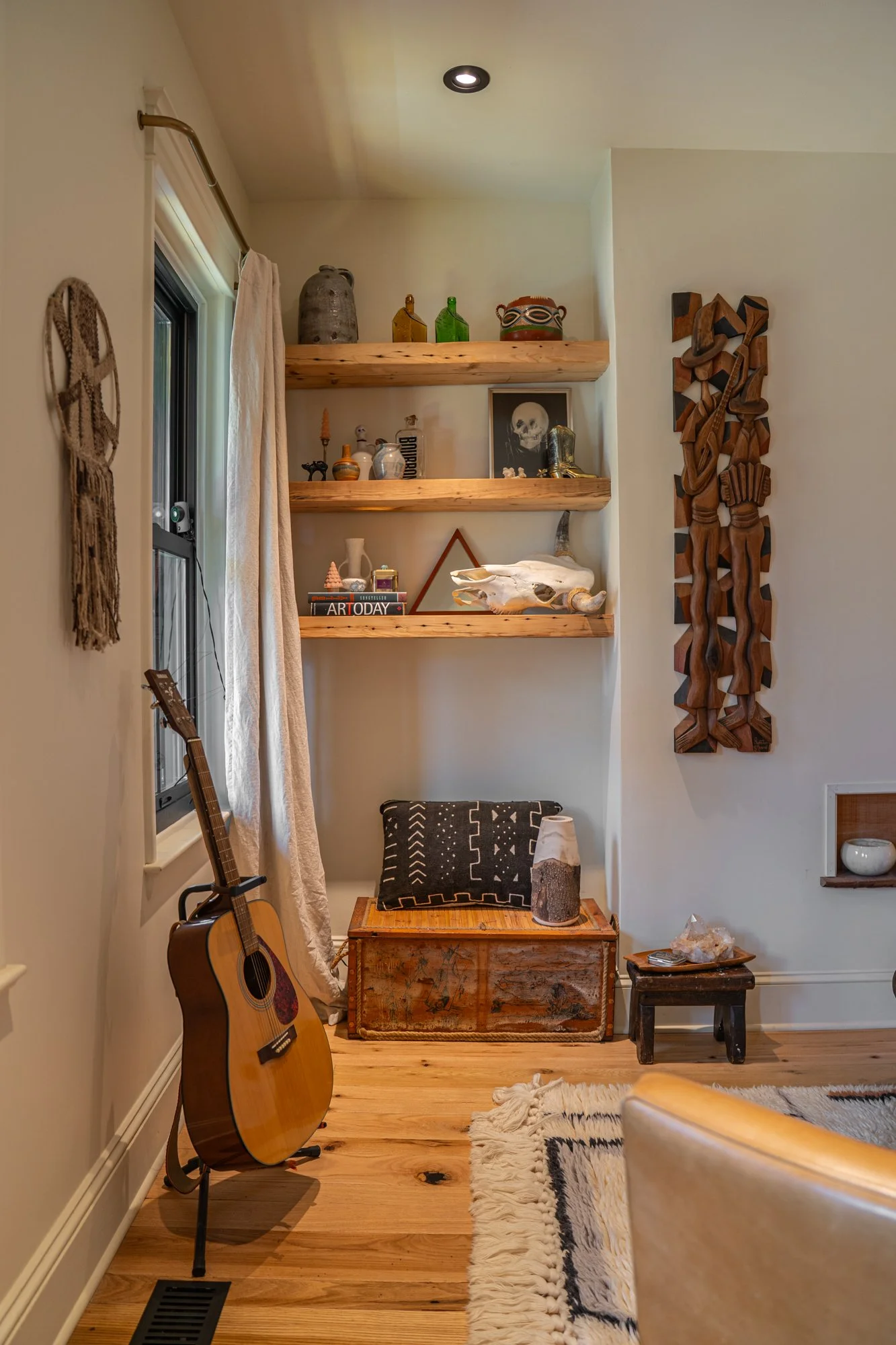A cozy corner of a room with a guitar on a stand next to a window, wooden shelves with decorative items, artwork, and a skull, a wooden chest with a pillow and vase, and a small side table on a wooden floor with a rug and a part of a chair.
