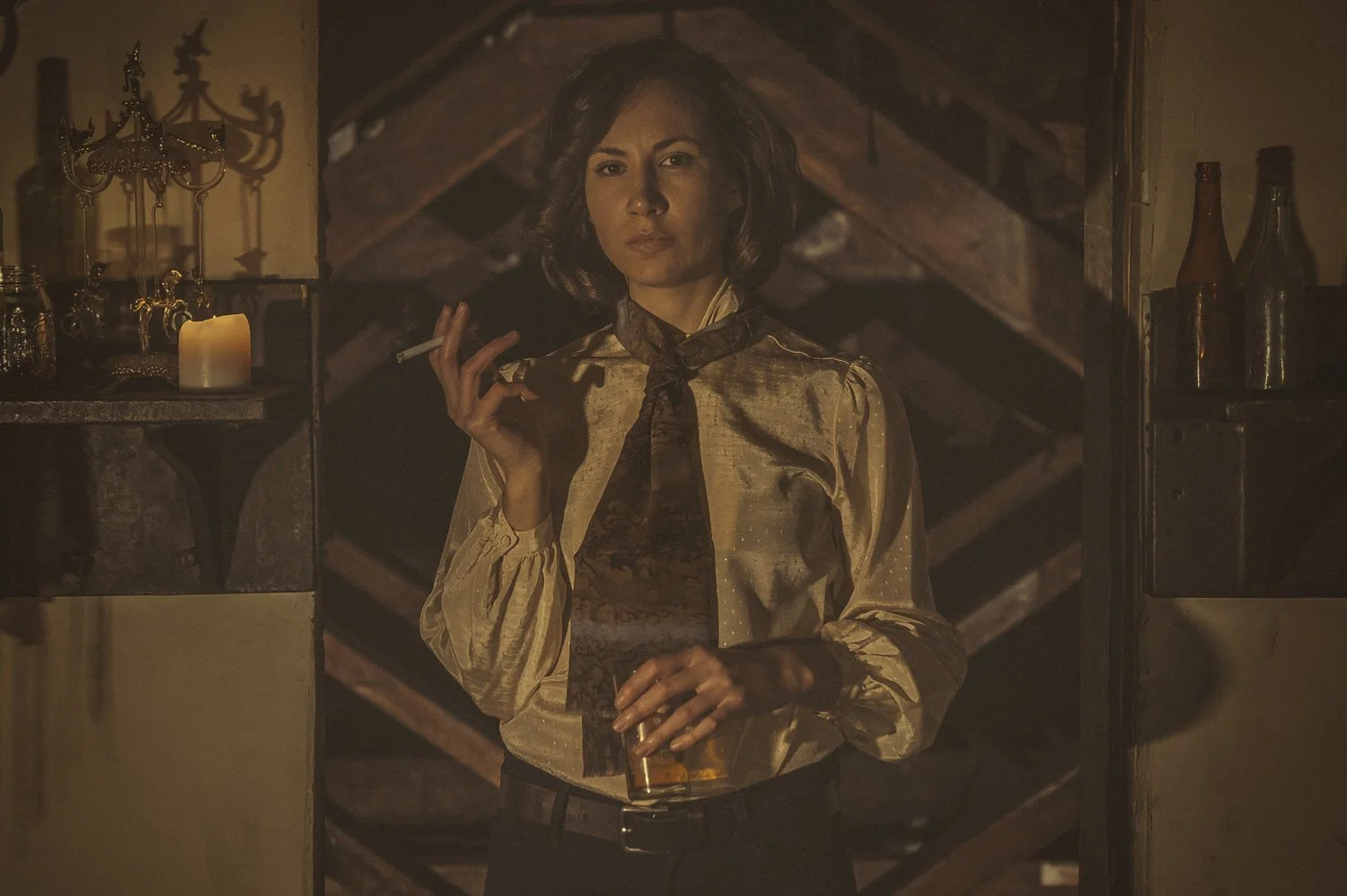 A woman standing in a dimly lit room, holding a cigarette in one hand and a glass of whiskey in the other, wearing a shiny beige blouse with a brown scarf.