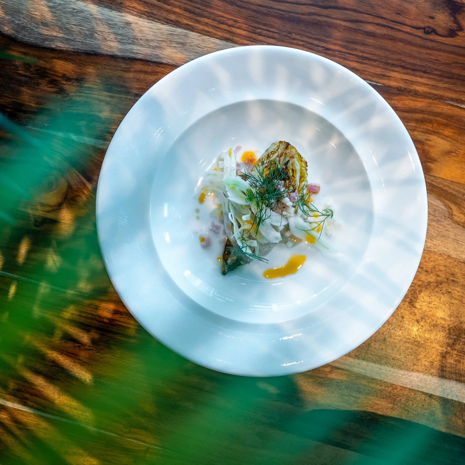 A gourmet dish featuring grilled vegetables and herbs on a white plate, with some green foliage in the foreground and a wooden table underneath.