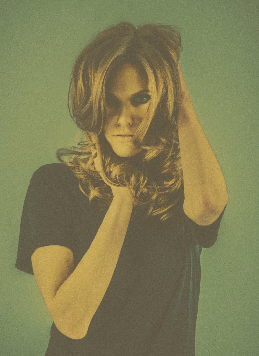A woman with long wavy hair, wearing a black top, posing with one hand on her head against a gradient green background.