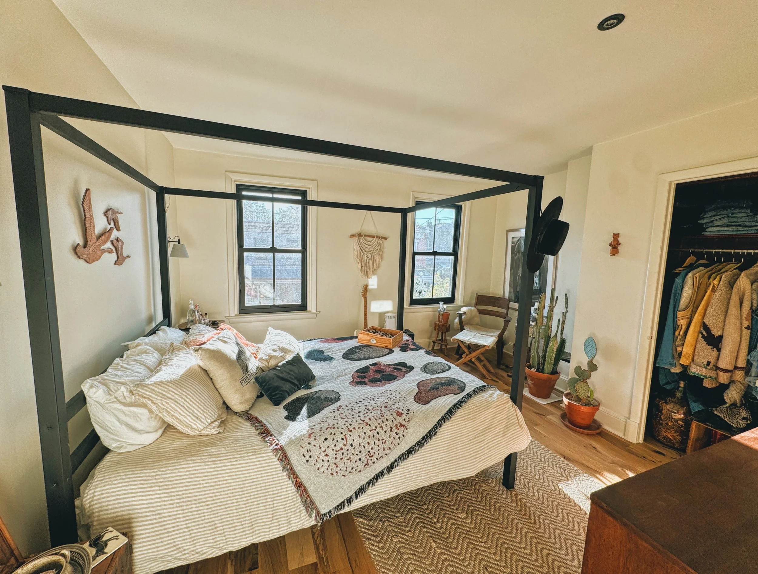 A bedroom with a black metal bed frame, a multi-colored throw blanket, multiple pillows, two windows, a small wooden chair, a large potted cactus, and an open closet with hanging clothes.