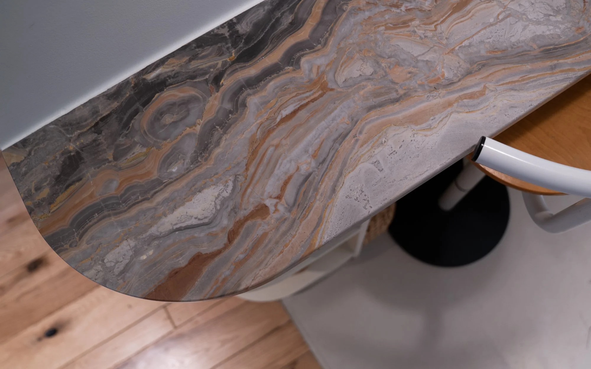 A marble countertop with swirling patterns in shades of gray, brown, and beige, next to a white wall and a wooden chair with white legs nearby.