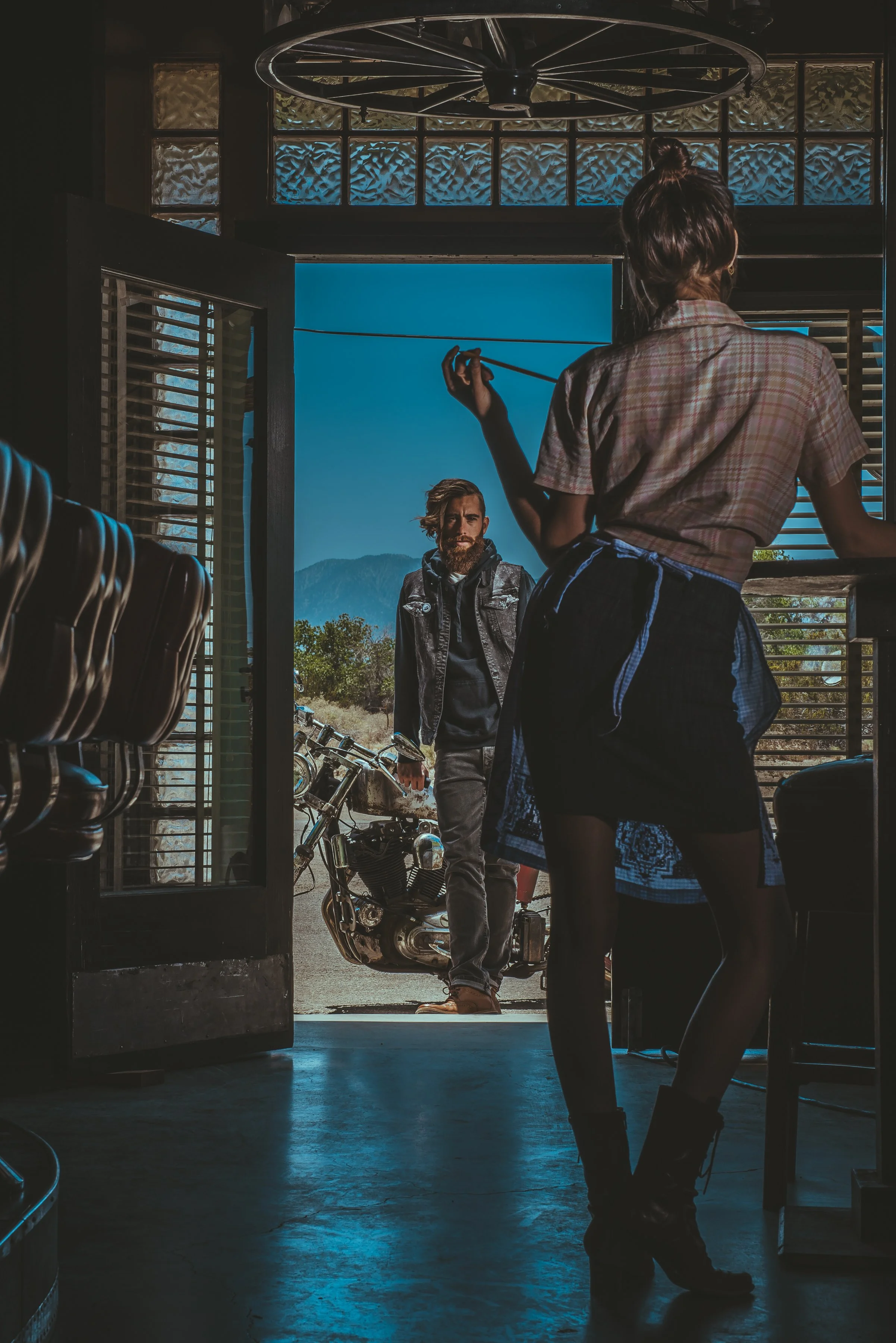 A woman with a plaid shirt and black skirt standing inside a shop near the counter, looking at a man outside. The man has a beard and long hair, wearing a leather vest and standing beside a motorcycle outside, with mountains in the background.