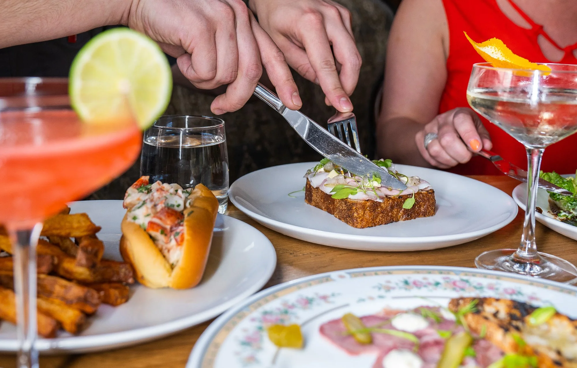 People dining at a restaurant with plates of food including a hot dog, fries, and a slice of bread topped with garnishes; drinks like a cocktail with a lemon slice, a glass of water, and a martini with an orange peel