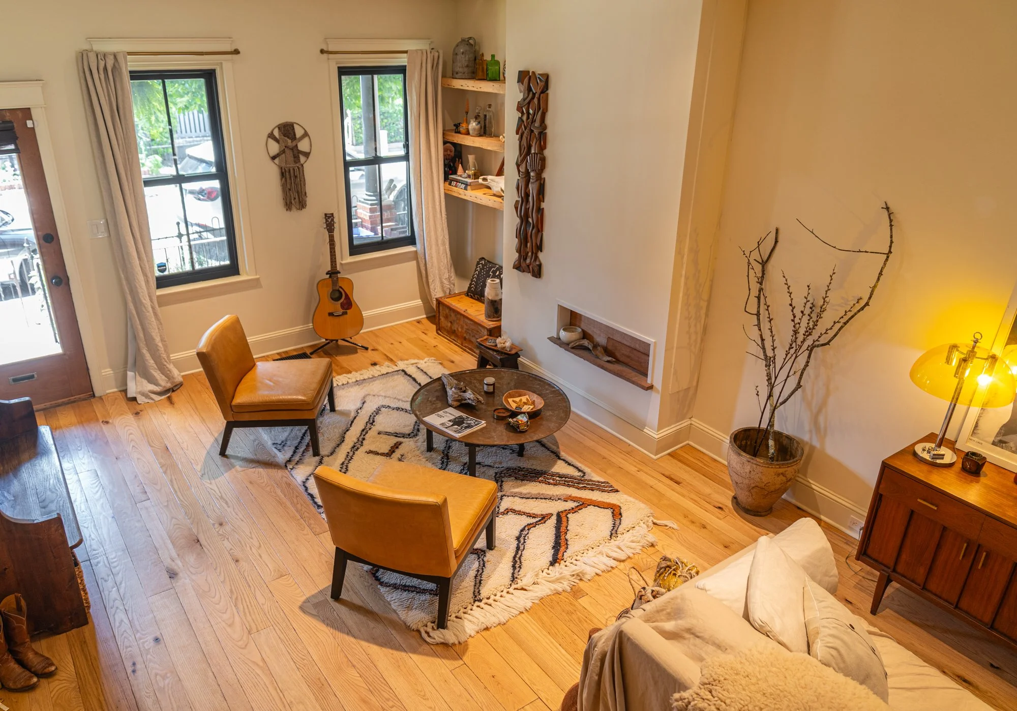 A cozy living room with hardwood floors, a patterned area rug, two yellow armchairs, a wooden coffee table with decorative items, a guitar near the window, and a wall shelf with decorative objects. There is a large potted branch in the corner, a side