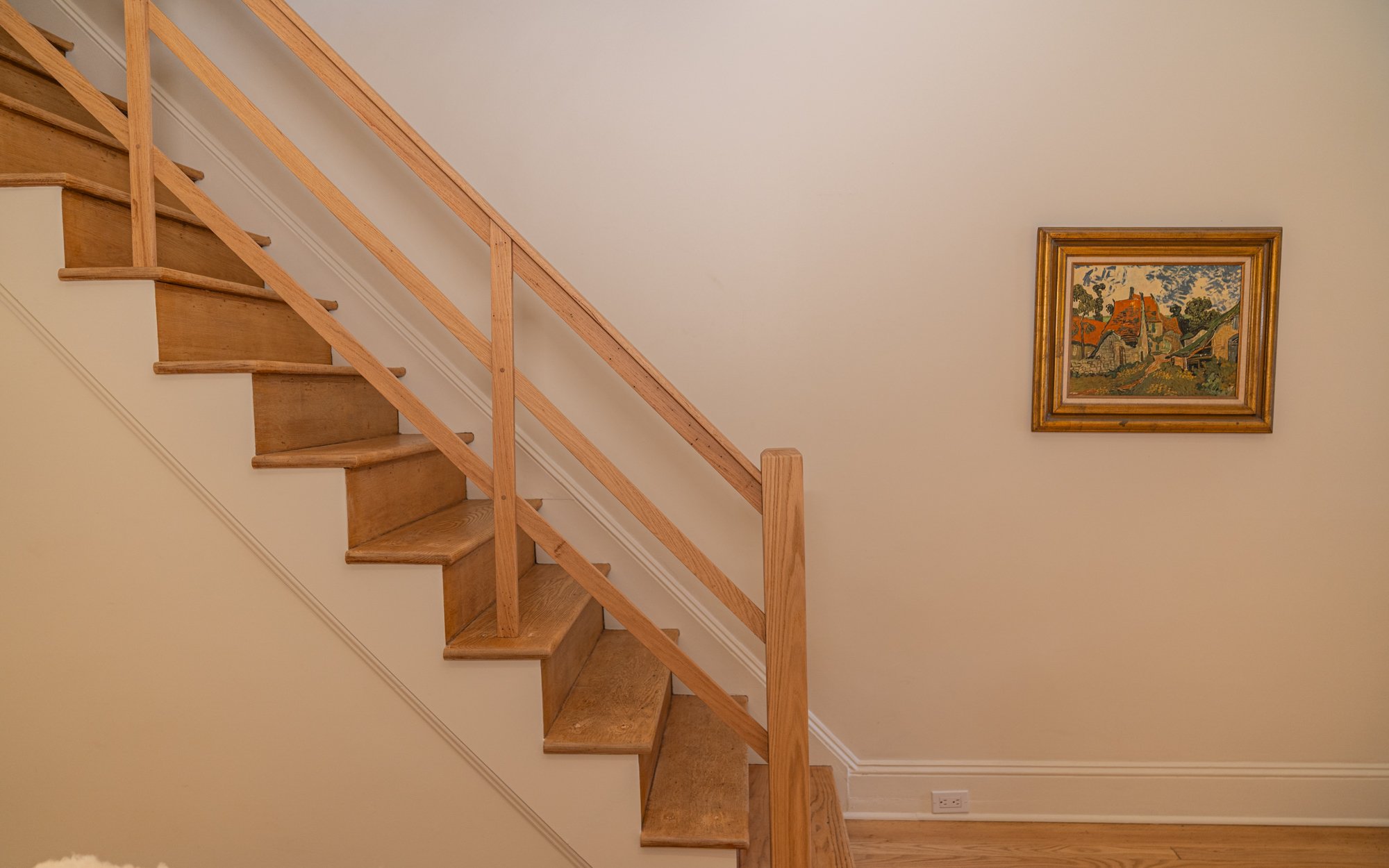 Interior of a house showing a wooden staircase with a handrail on the left and a framed landscape painting on the right wall.