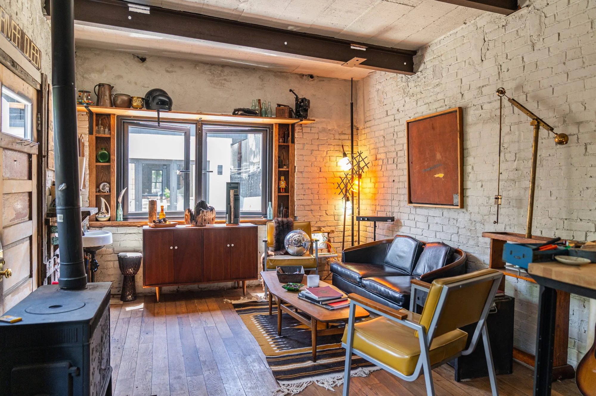 An eclectic living room with white brick walls, wooden flooring, and modern furniture. Includes a black leather sofa, yellow armchair, coffee table with books, and a large window with decorative objects on the sill. Decorated with retro and vintage i