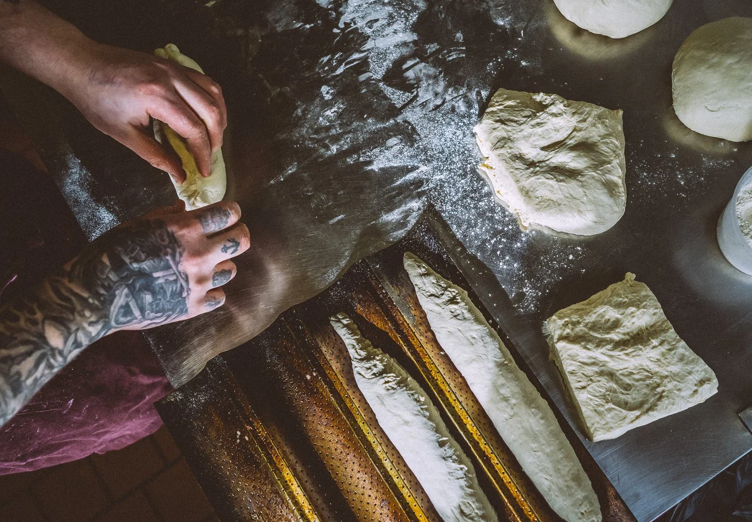 A person with tattoos stretching dough on a floured surface, with several pieces of shaped dough ready for baking.