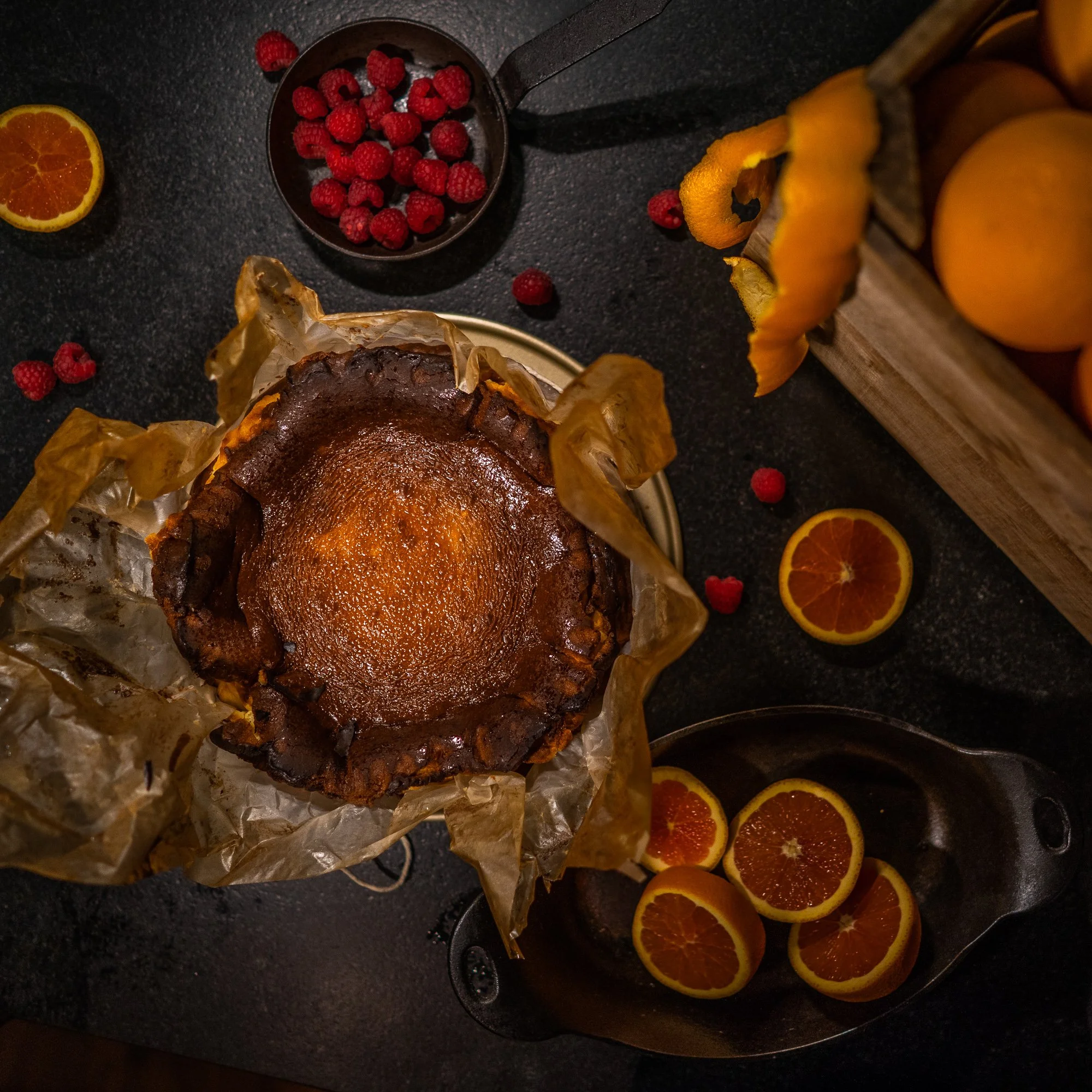 A baked pie with a golden, crispy crust on a dark countertop, surrounded by halved oranges, a small bowl of raspberries, and a wooden crate filled with oranges.