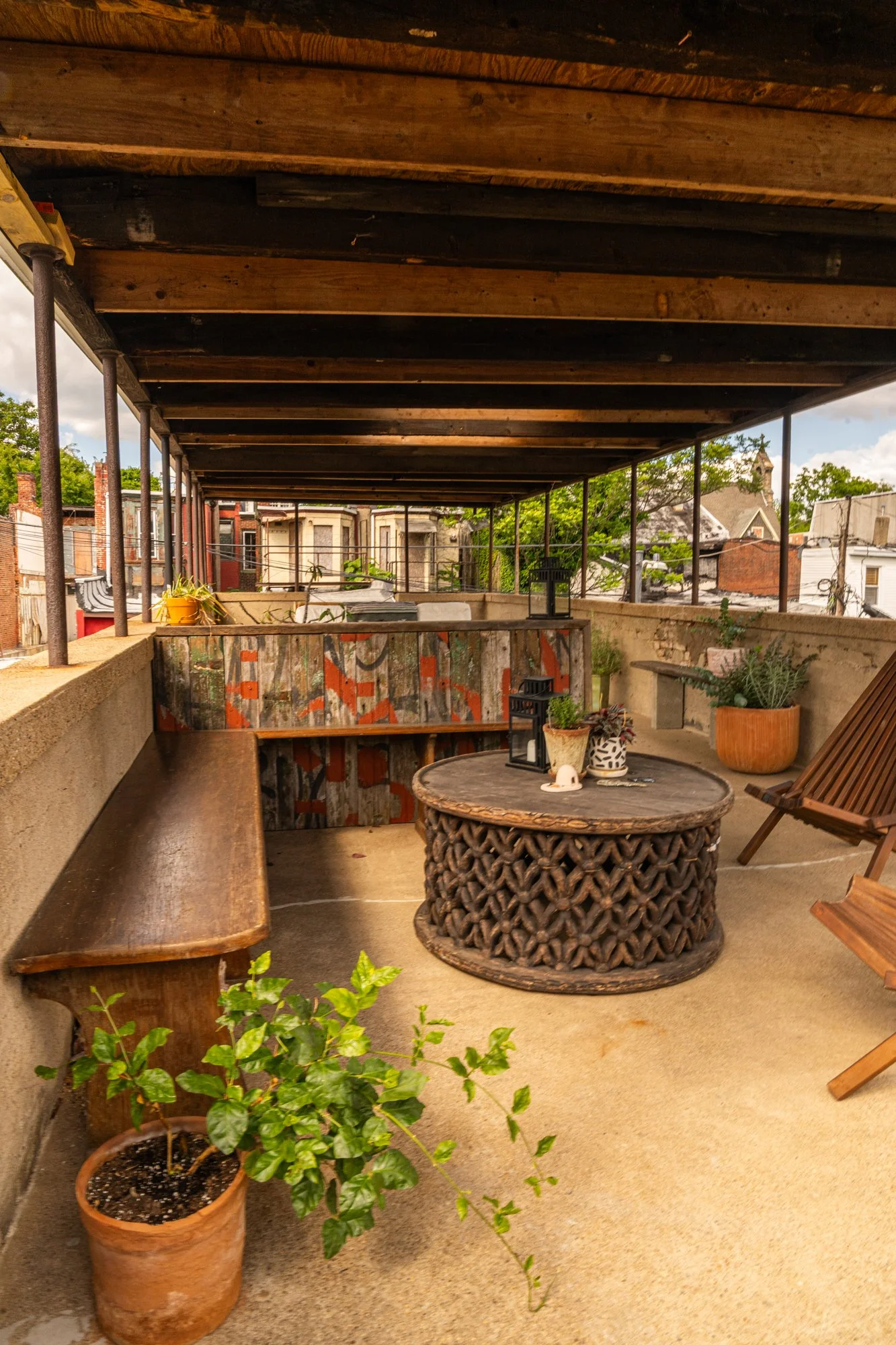 A rooftop patio with a wooden bench, a round woven coffee table, plants, and lanterns. The patio has a low wall with painted graffiti and a protective roof structure. Residential buildings are visible in the background.