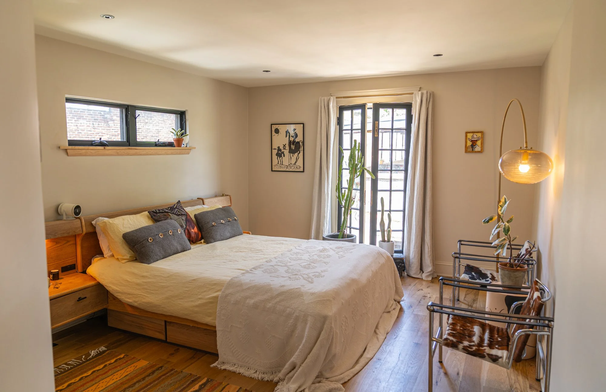 A cozy bedroom with a wooden bed, decorative pillows, and natural light from large windows and a door with curtains. There are potted cacti near the door, framed artwork on the walls, and a leopard print chair with a nearby floor lamp.