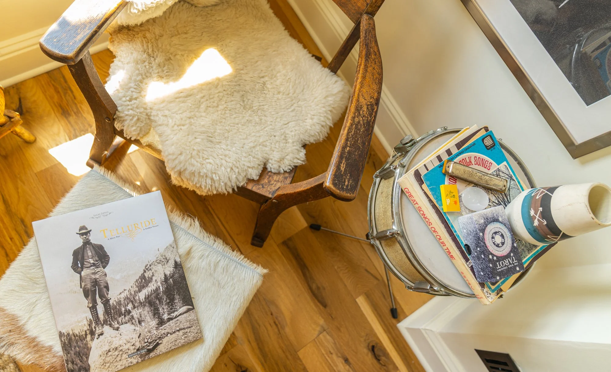 An overhead view of a corner with a wooden chair with a sheepskin seat, a stack of vintage magazines on a small round table, and a book titled 'Telluride' on a furry white cushion. The magazines include music and art covers, and there is a white mug 