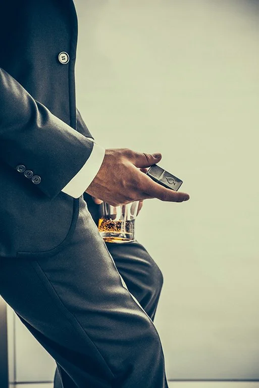 A person in a gray suit sitting down, holding a remote control, with a glass of whiskey on a table nearby.