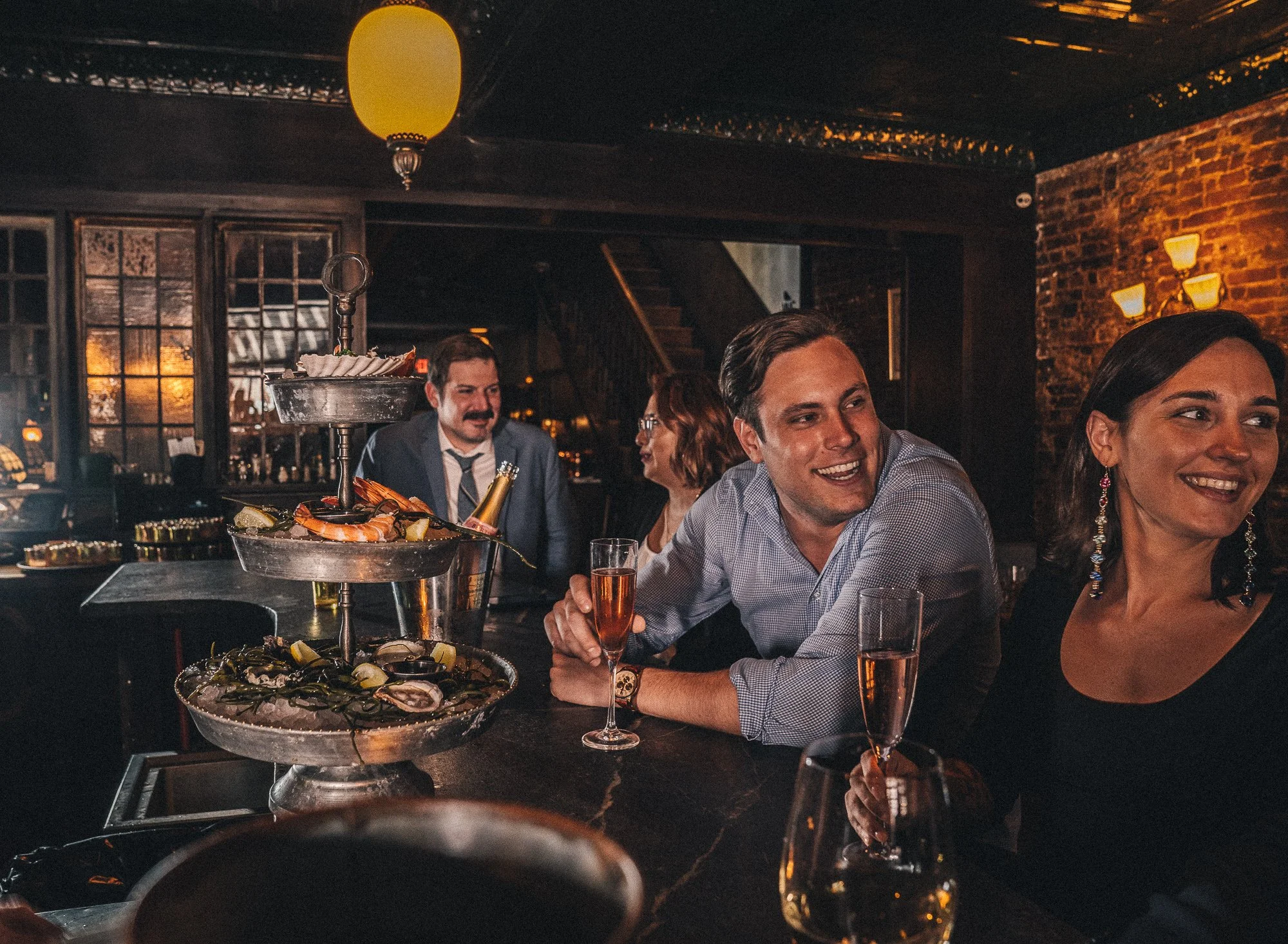 People enjoying drinks and conversation at a bar with seafood on a tiered platter in front of them.