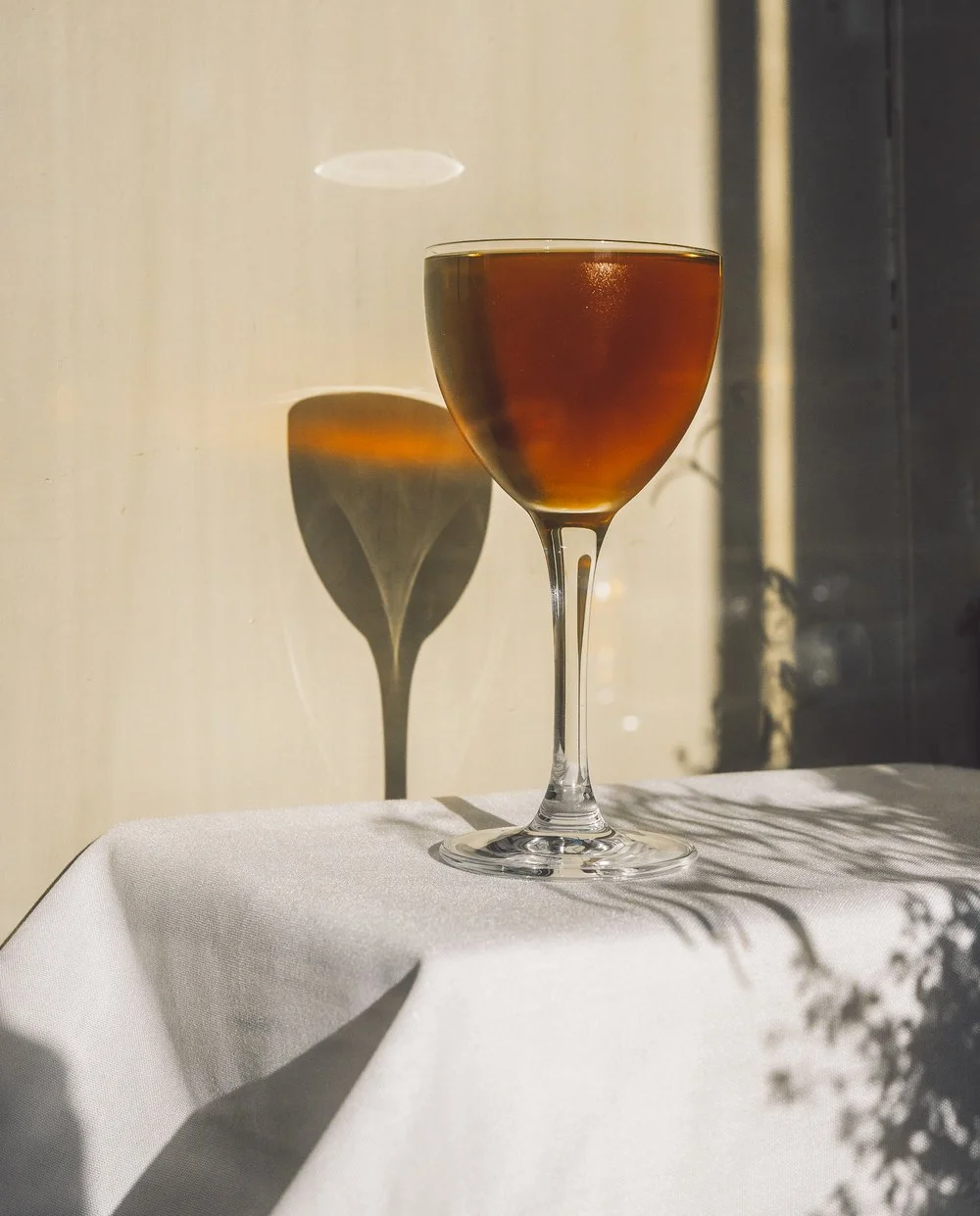 A glass of dark amber liquid on a white tablecloth with shadows cast on the wall behind it.