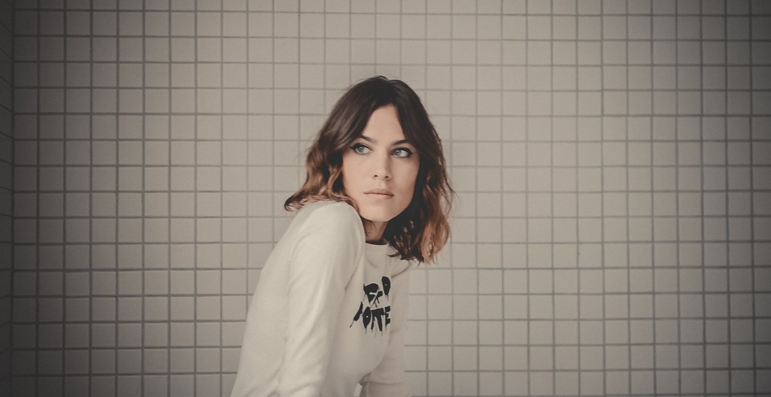 A young woman with shoulder-length wavy brown hair and blue eyes looking to her right, wearing a white sweatshirt with black text, against a tiled wall background.