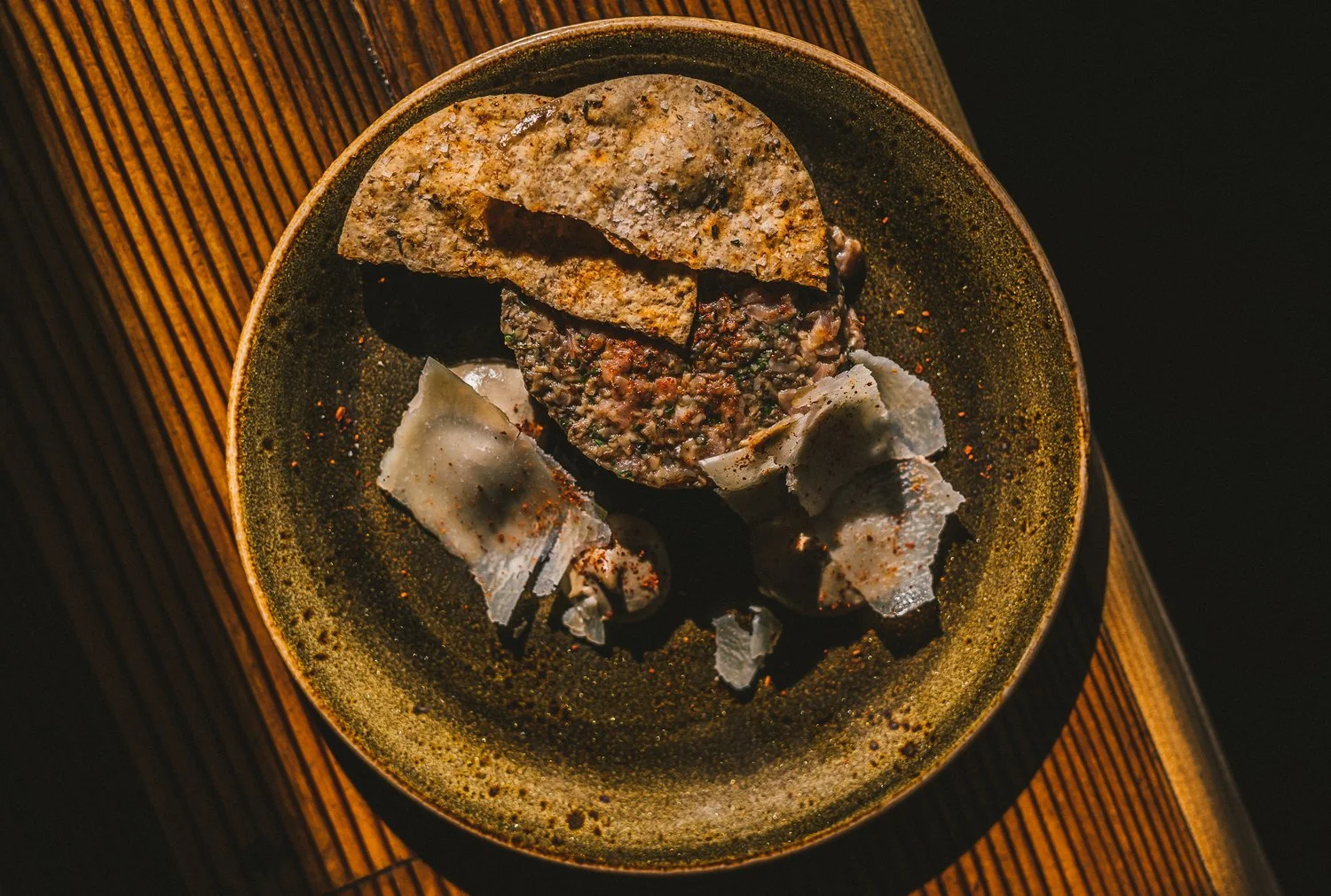 A ceramic bowl containing a patty and broken pieces of flatbread or chips, placed on a wooden surface.