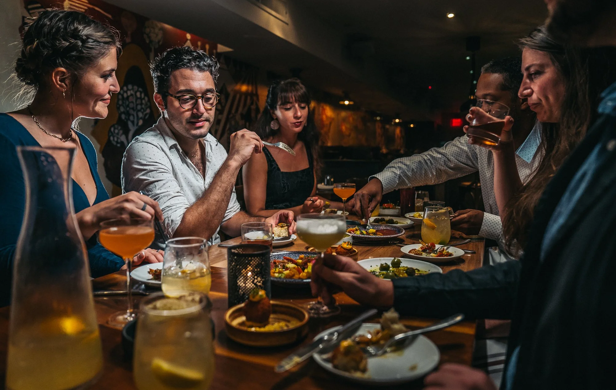 Group of six friends enjoying a dinner gathering at a restaurant, with various plates of food and drinks on the table, warmly lit in a cozy setting.