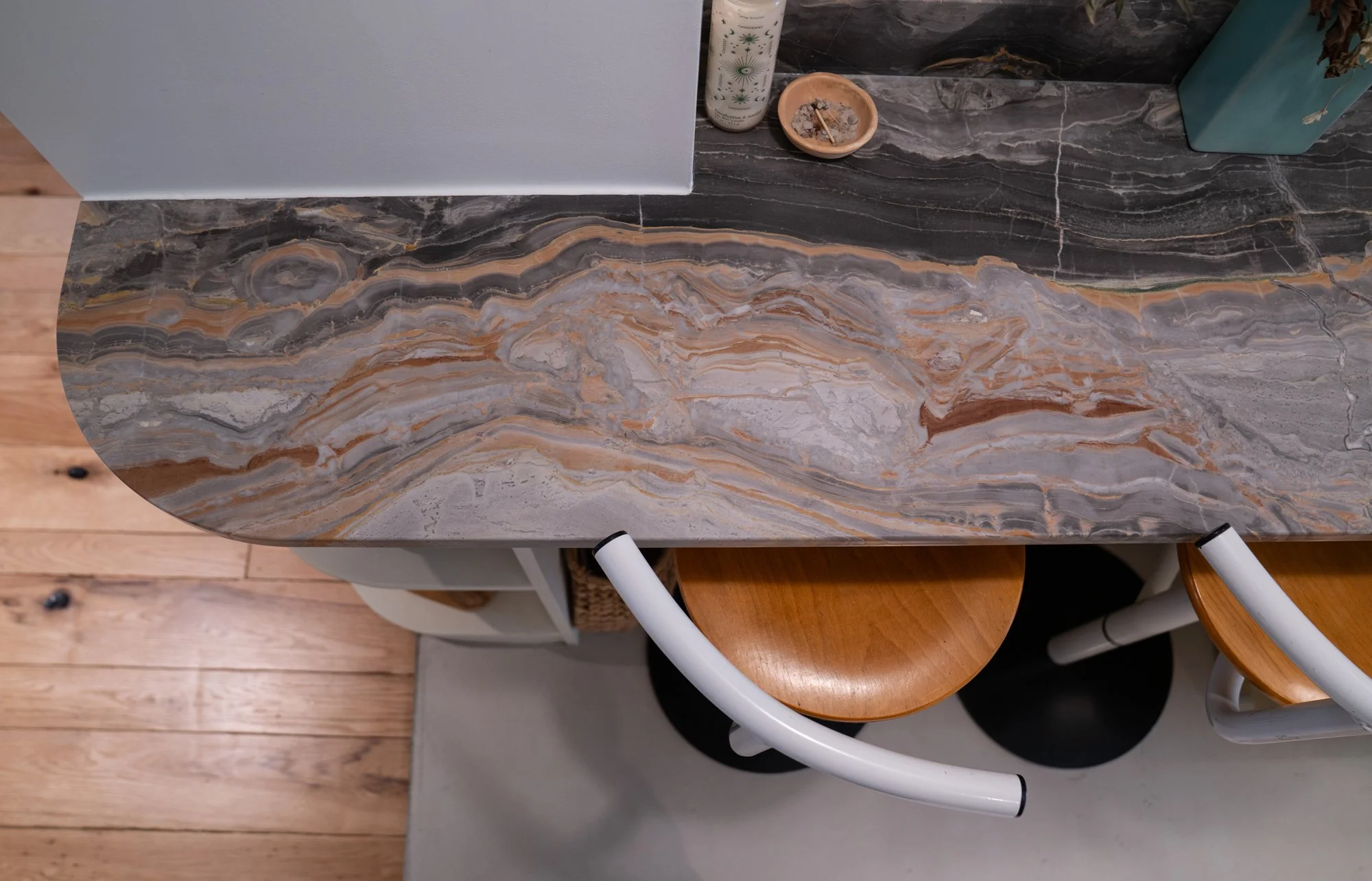 Top view of a marble countertop with shades of gray, brown, and white, with two wooden chairs with curved white backs tucked underneath. The floor is wooden with visible black knots.