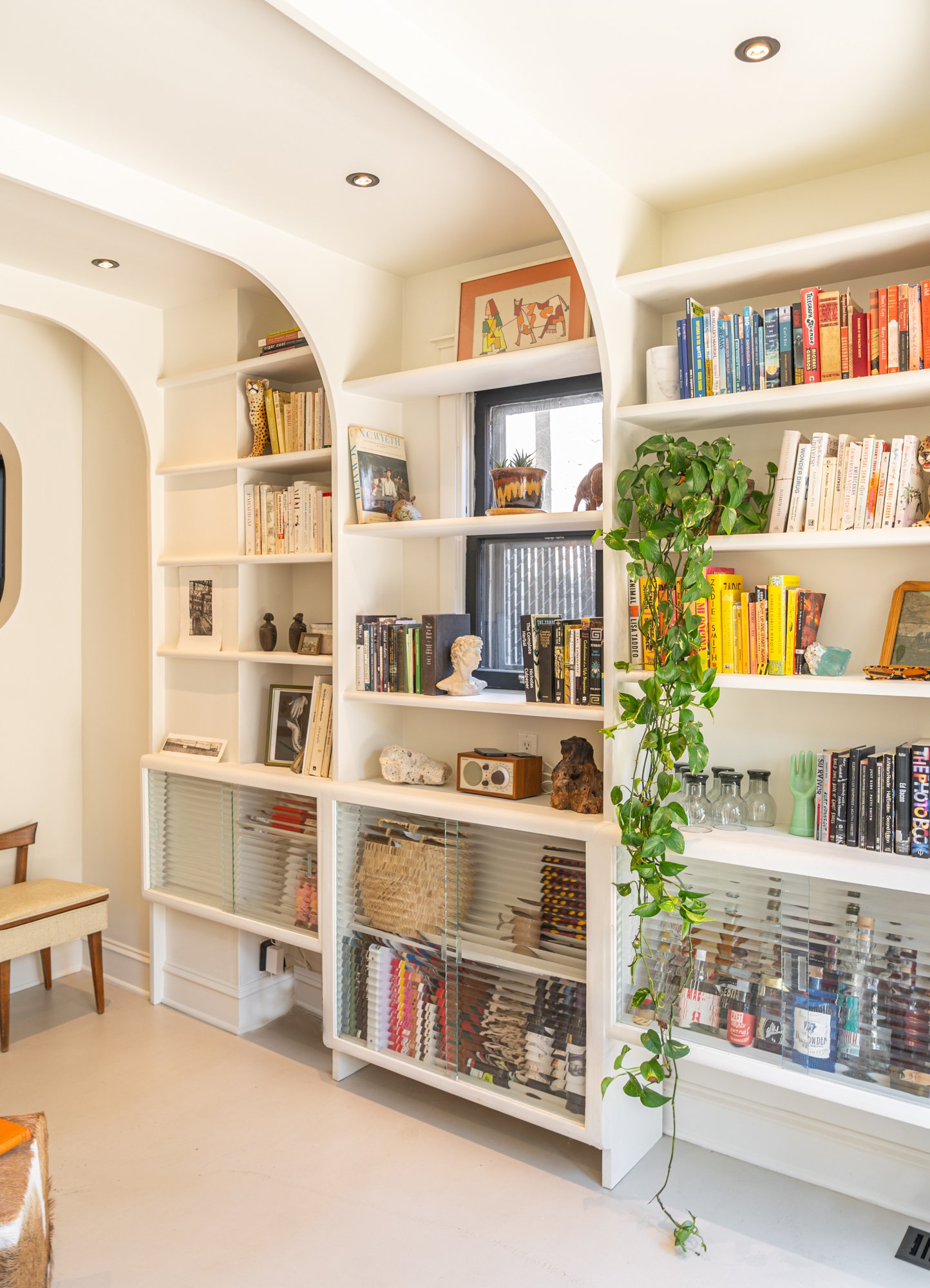 A white built-in bookshelf filled with books, decorative objects, and artwork in a room with beige carpet and a small window with black trim.